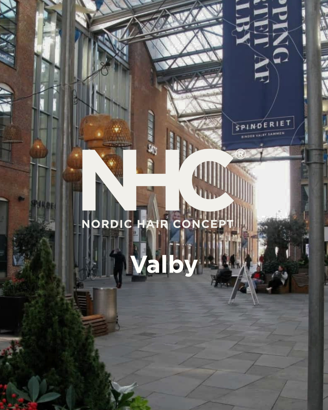Nordic Hair Concept Valby i Spinderiet. Uden for en shoppinggade i Valby, København, med trafikkegler, træer, butikker og folk, der går og sidder på bænke.
