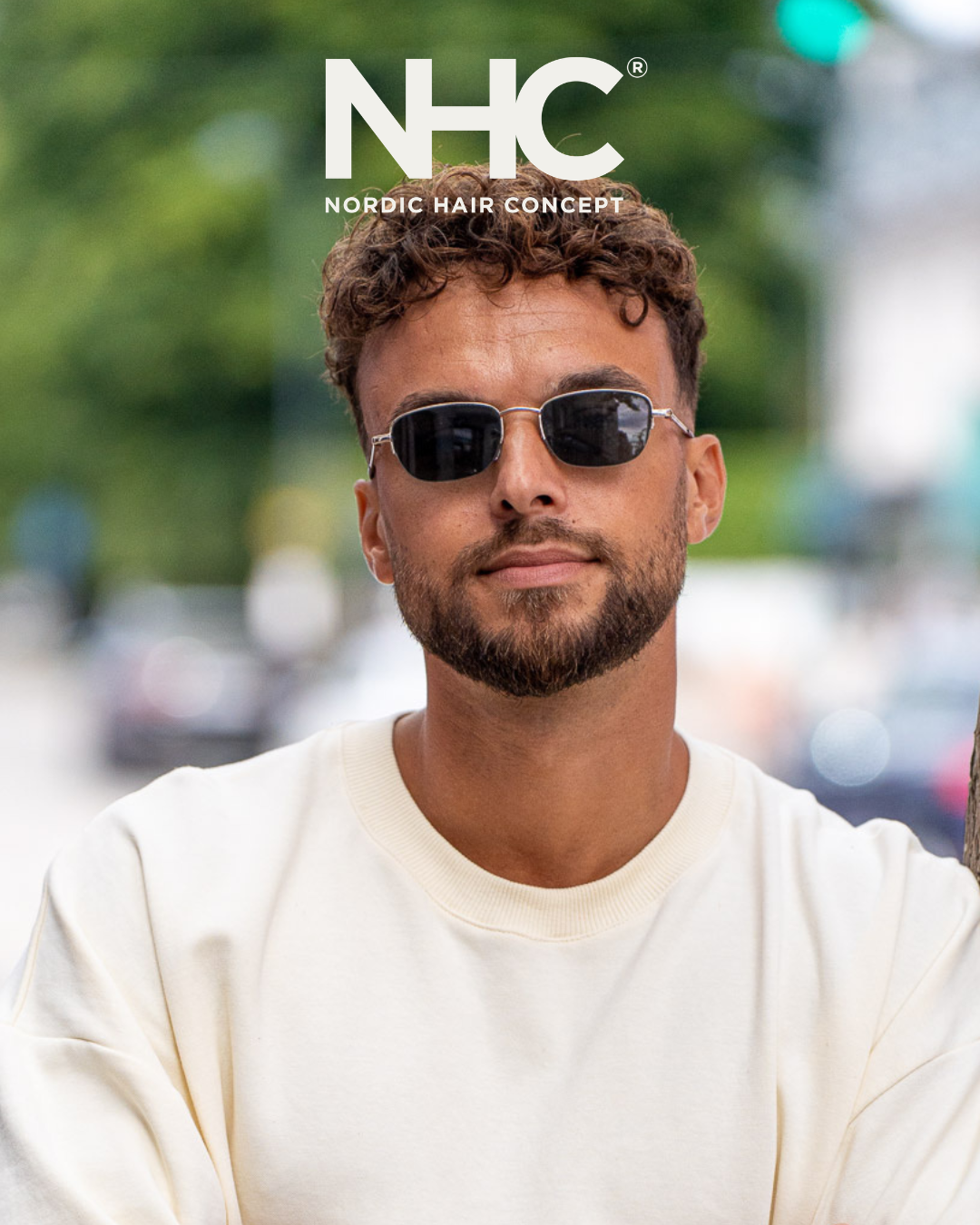 Mand med skæg, mørke solbriller og lyst sweatshirt står udenfor med grønne træer i baggrunden.