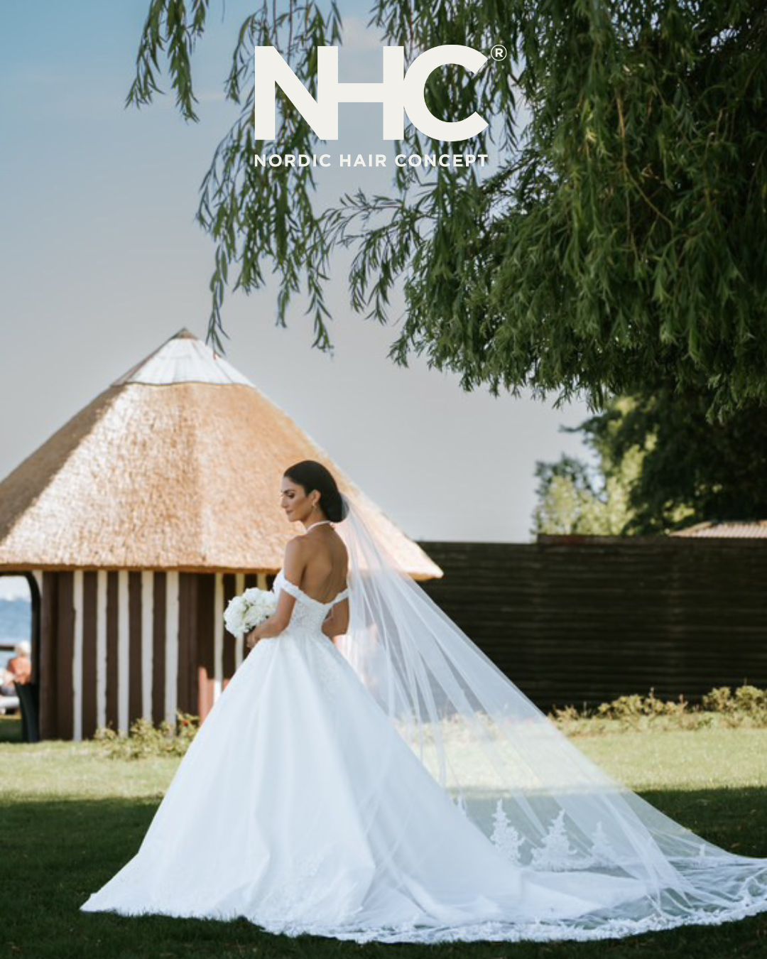 En brud i hvid brudekjole med lang slør står udenfor under træer, med en hytte i baggrunden.