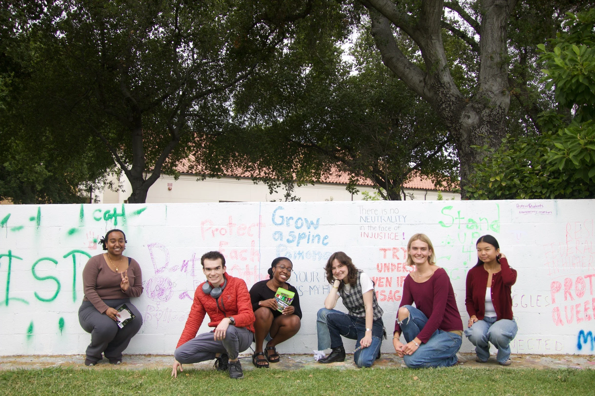 Group of six diverse young people smiling and kneeling in front of a white wall with colorful graffiti and messages, under large trees on a grassy area.