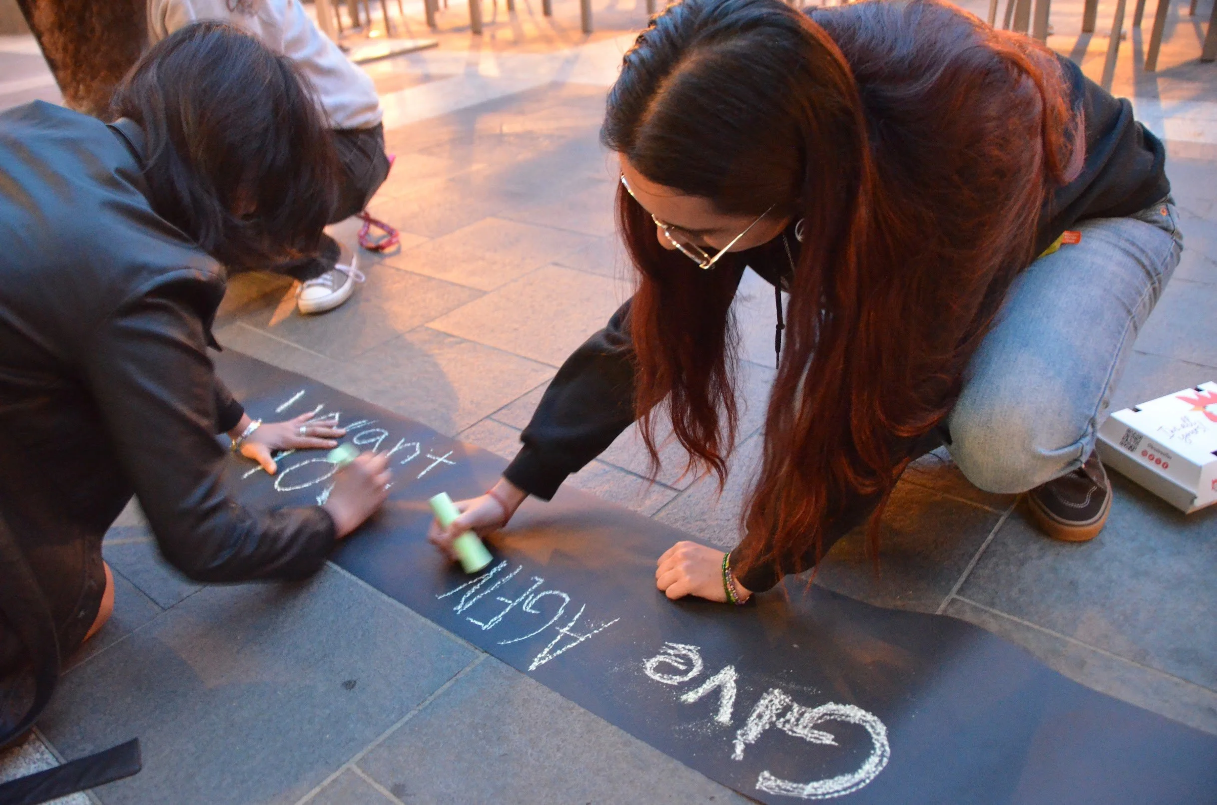 Two people are kneeling on the sidewalk, writing messages with chalk on a black strip of paper that is laid on the ground. One person is a woman with long red hair, glasses, and casual clothes. The other person is a girl with long dark hair, wearing a black jacket.
