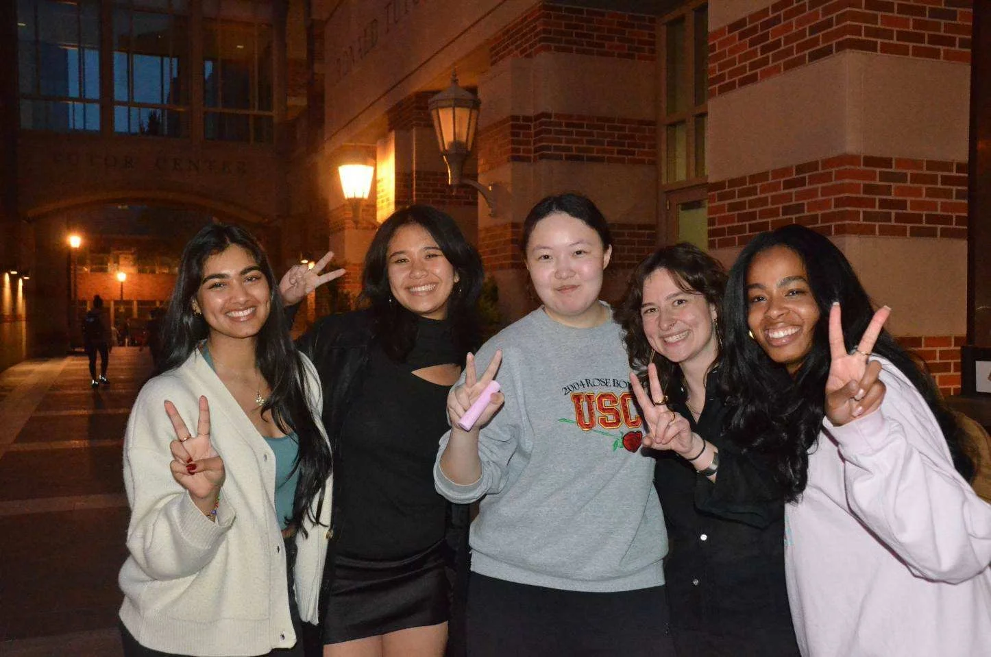 A group of five young college women smiling and making peace signs with their hands, standing outside at night in front of a brick building with streetlights.