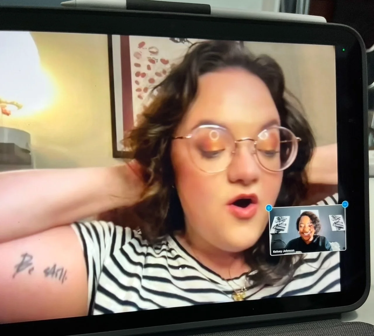 A woman with wavy dark hair wearing large round glasses and a striped shirt, with her eyes closed and mouth slightly open, in a video call with another woman who has curly hair and is smiling.