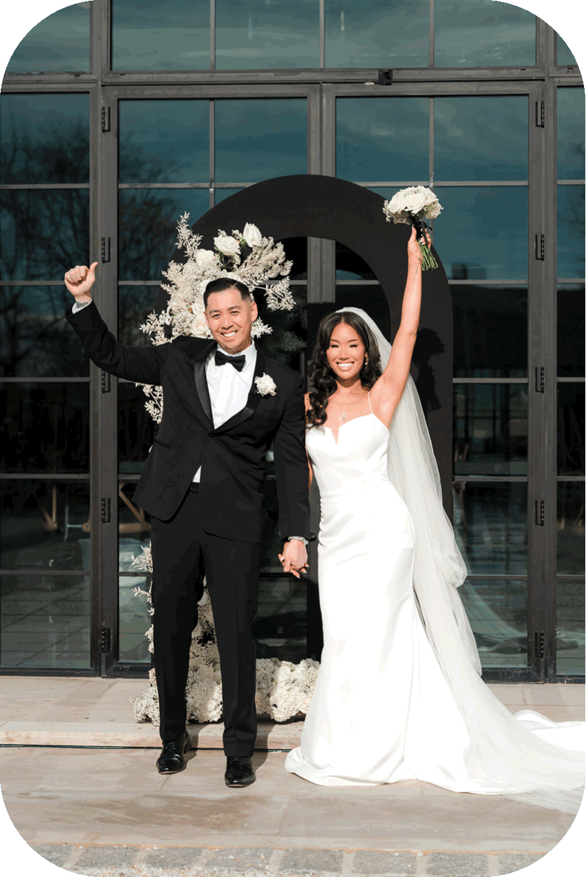 A bride and groom celebrating at their wedding, holding hands and smiling, with the bride raising a bouquet of white flowers in the air. They are standing in front of a decorative black circle with white floral arrangements, inside a building with large glass windows.