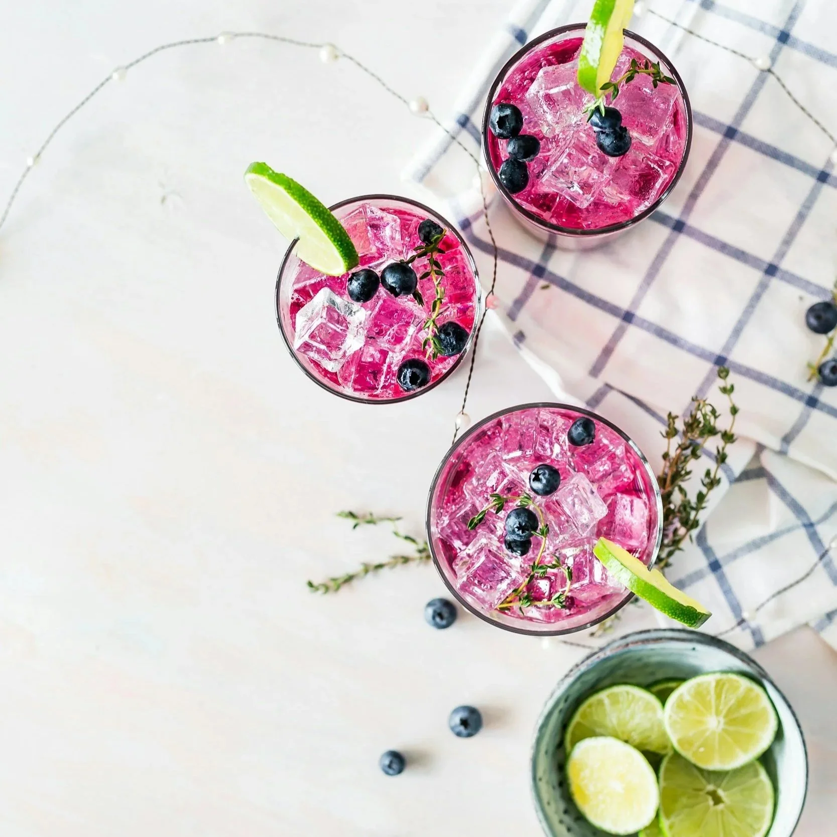 Four pink drinks with ice, blueberries, lime wedges, and herbs on a white surface, with a checkered cloth and a bowl of lemon slices nearby.
