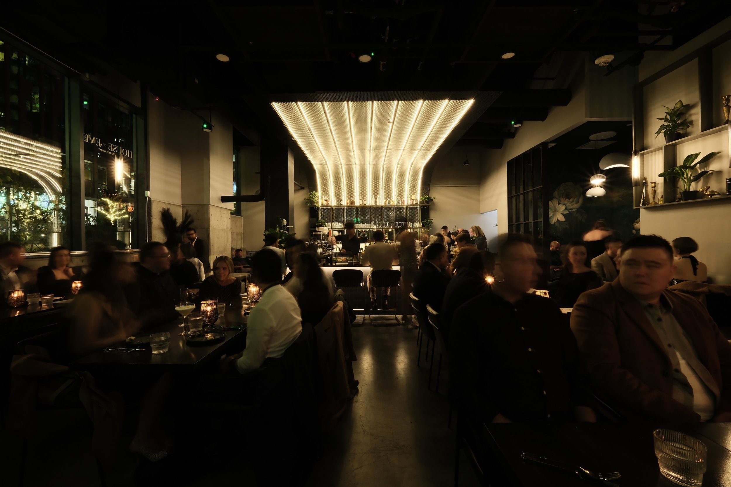 Dimly lit modern restaurant interior with patrons dining and a central illuminated bar.