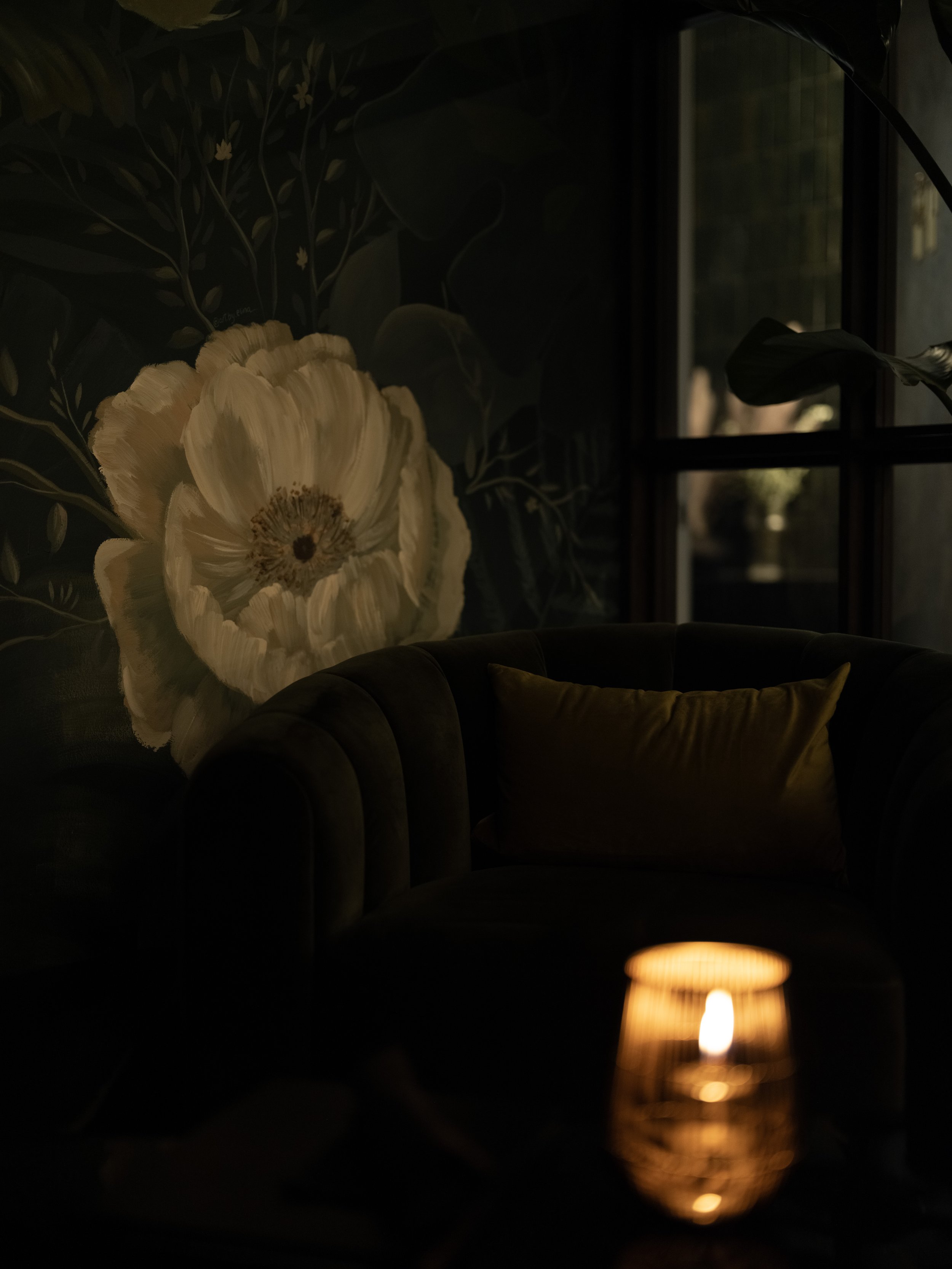 Dimly lit room with a candle on a table, a dark green velvet sofa, and a floral mural on the wall.