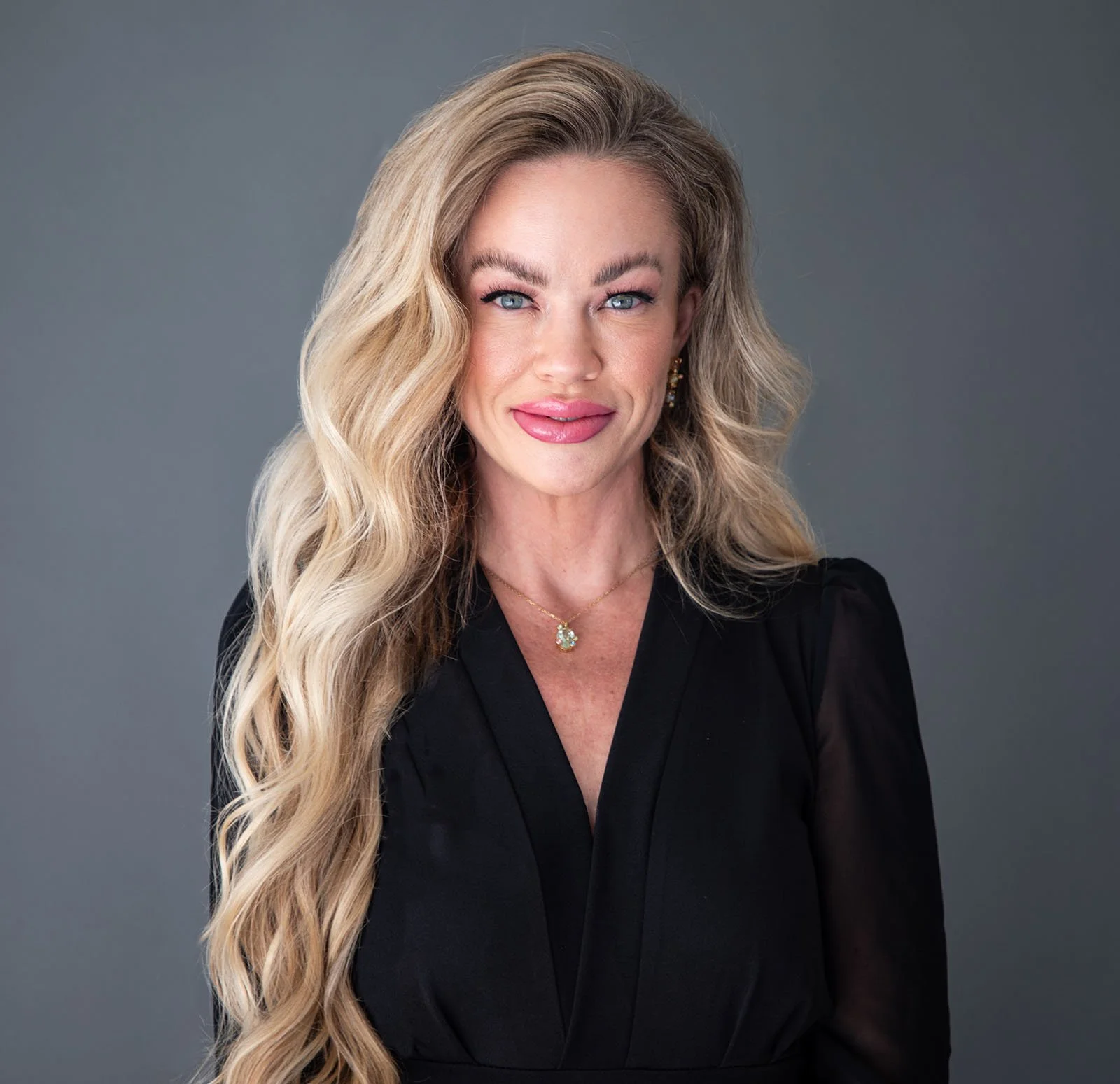 A woman with long, wavy blonde hair wearing a black top, gold earrings, and a gold necklace with a pendant, smiling and posing against a gray background.