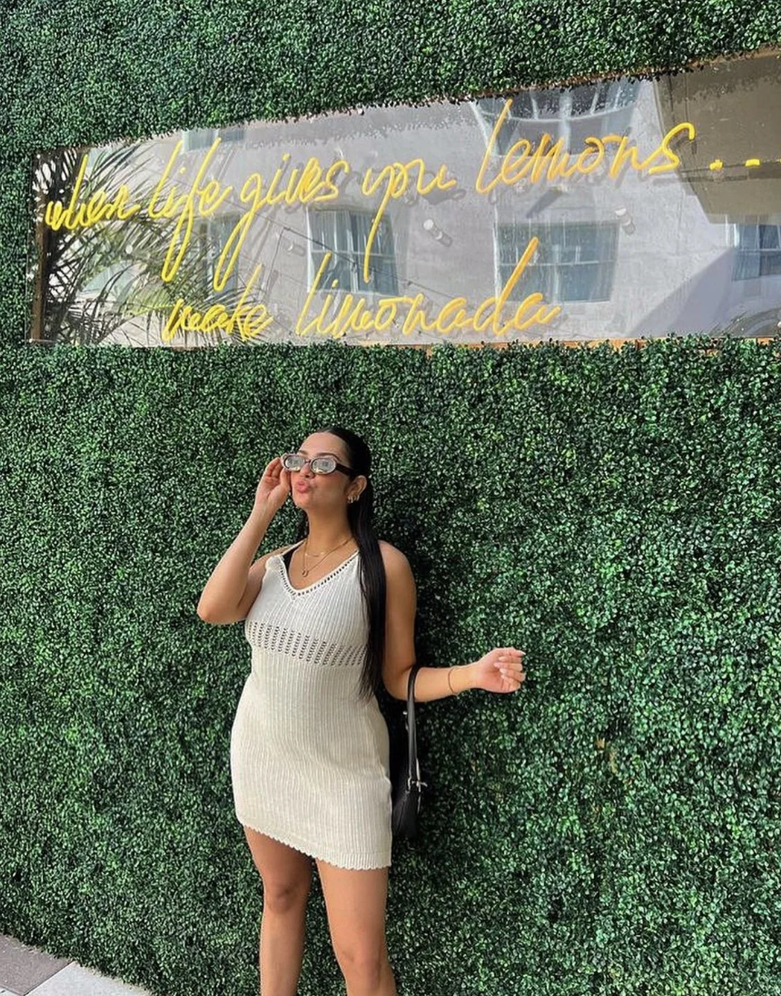 A woman in a white knitted dress and sunglasses posing in front of a green hedged wall with a neon sign that reads 'when life gives you lemons... make mojitos' and a reflection of buildings in a window above