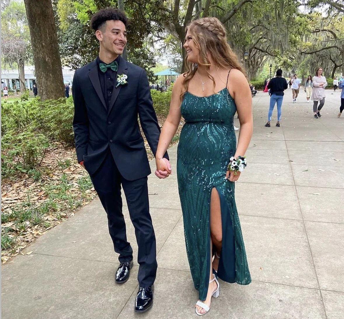 A young man in a black suit with a green bow tie and a woman in a teal sequined gown with a slit, holding hands and walking outdoors in a park with other people in the background.