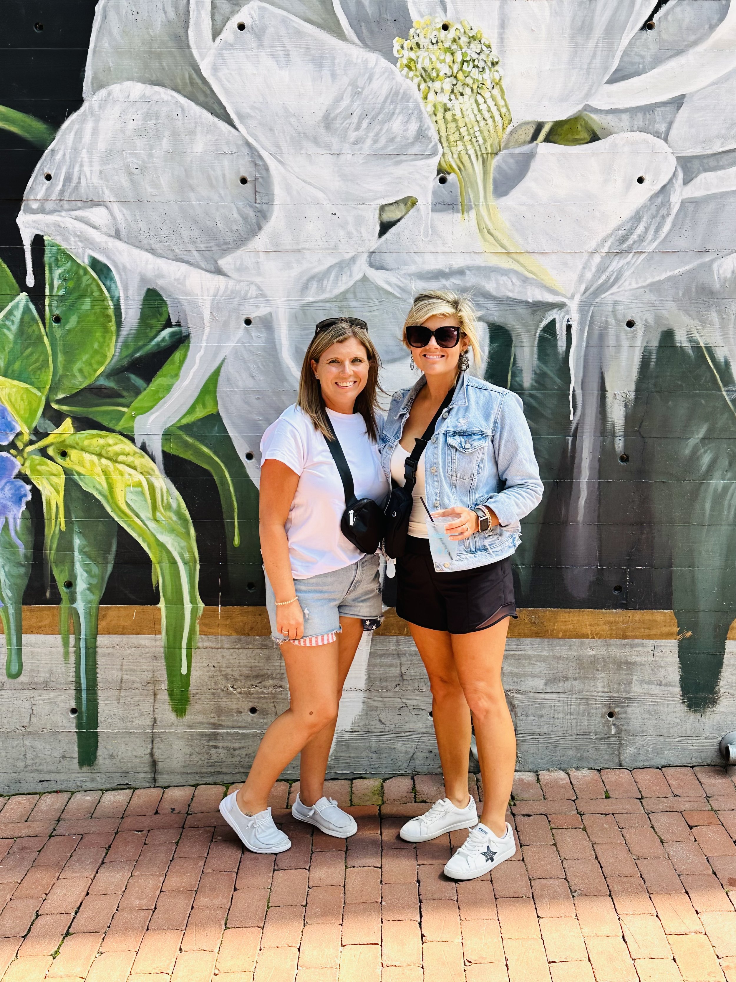 Two women standing in front of a colorful mural of a white flower with green leaves, on a brick-paved sidewalk.