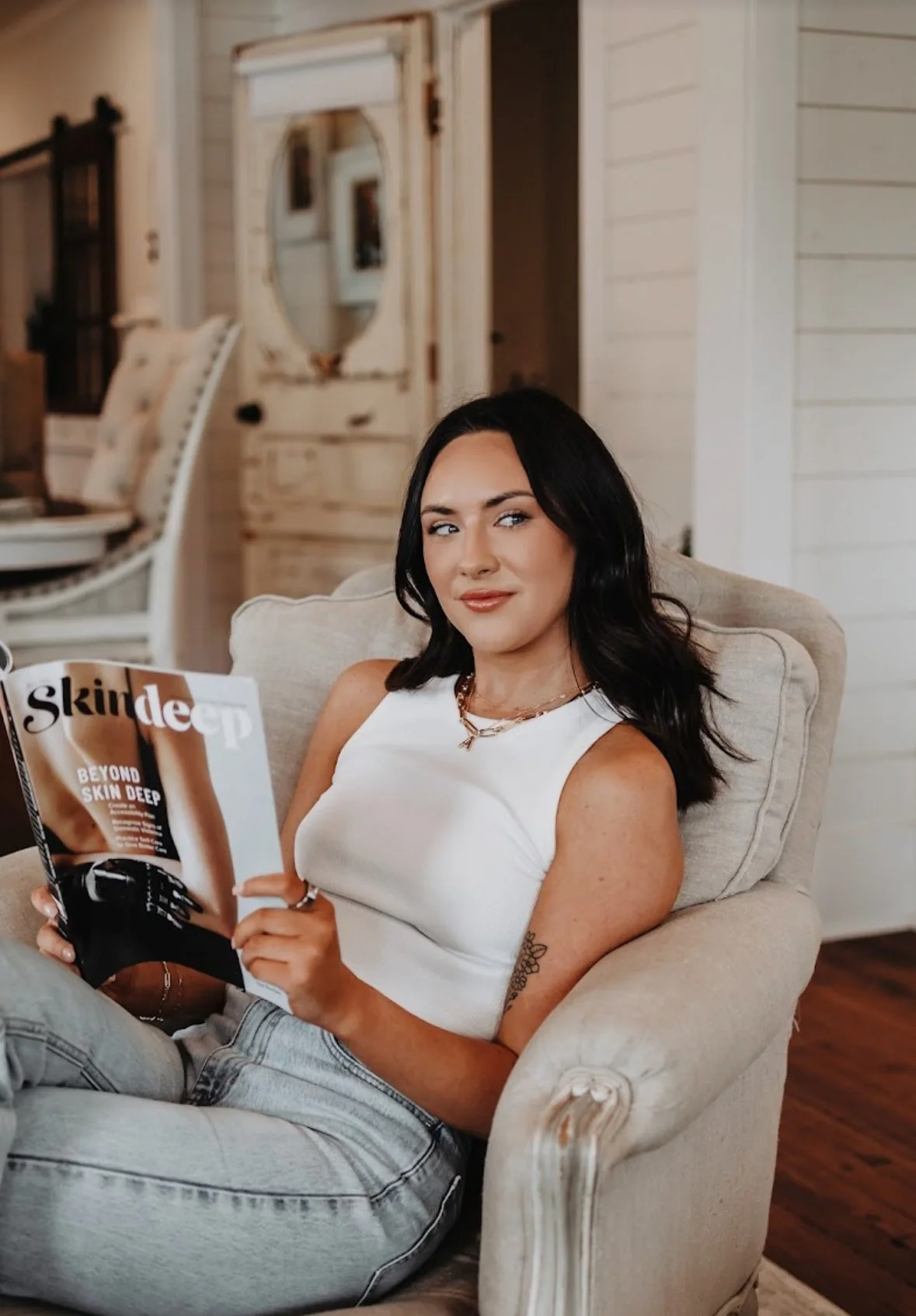 A woman with dark hair, wearing a white tank top, sitting on a beige armchair, holding a magazine and looking at the camera in a cozy, well-lit room.