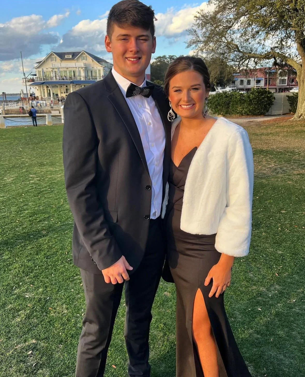 A young man and woman dressed in formal attire standing outdoors on grass, smiling at the camera, with waterfront and buildings in the background.