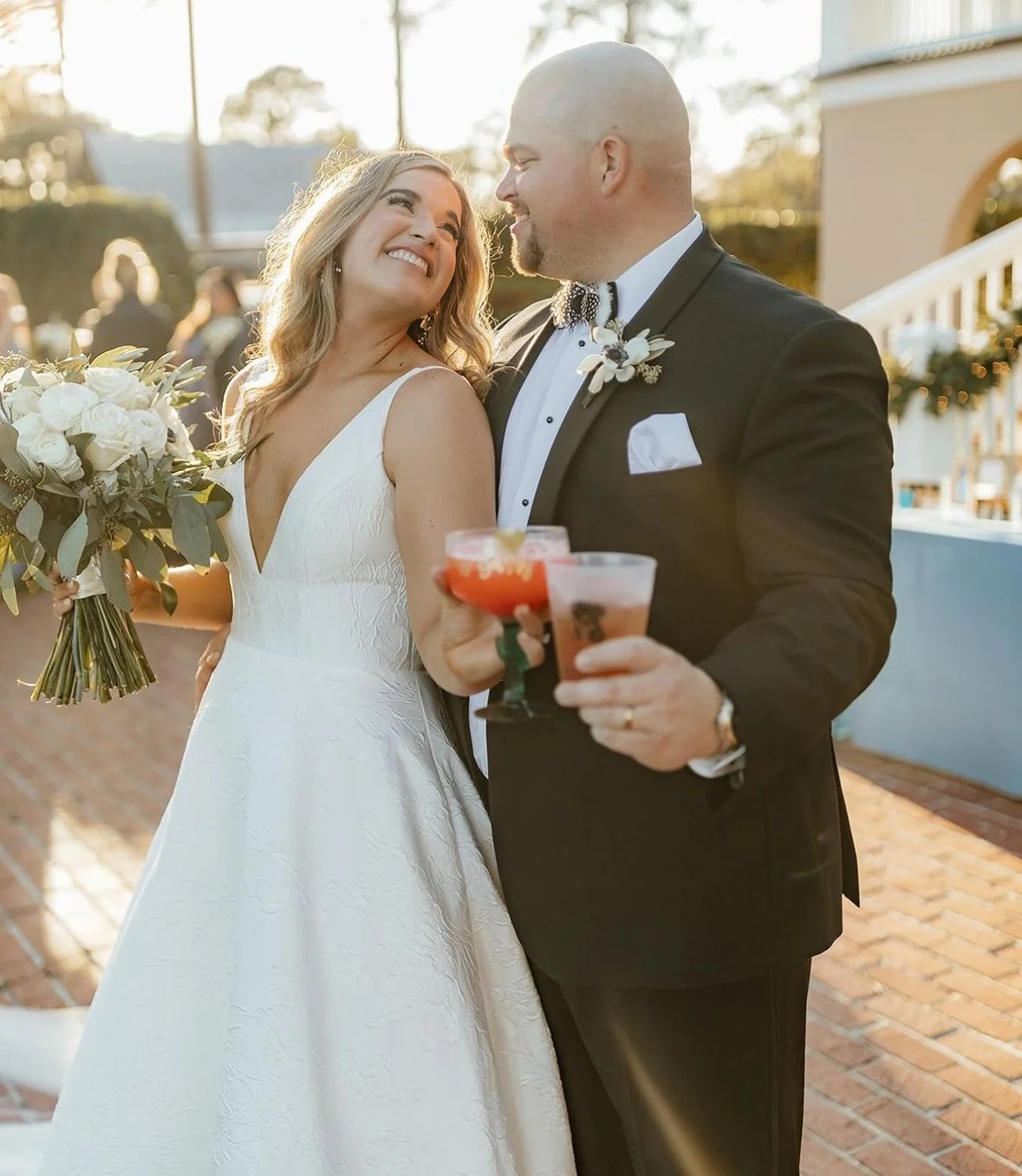 A newlywed couple at their wedding celebration, smiling and holding cocktails, with the bride holding a bouquet of white flowers and greenery, outdoors during sunset.