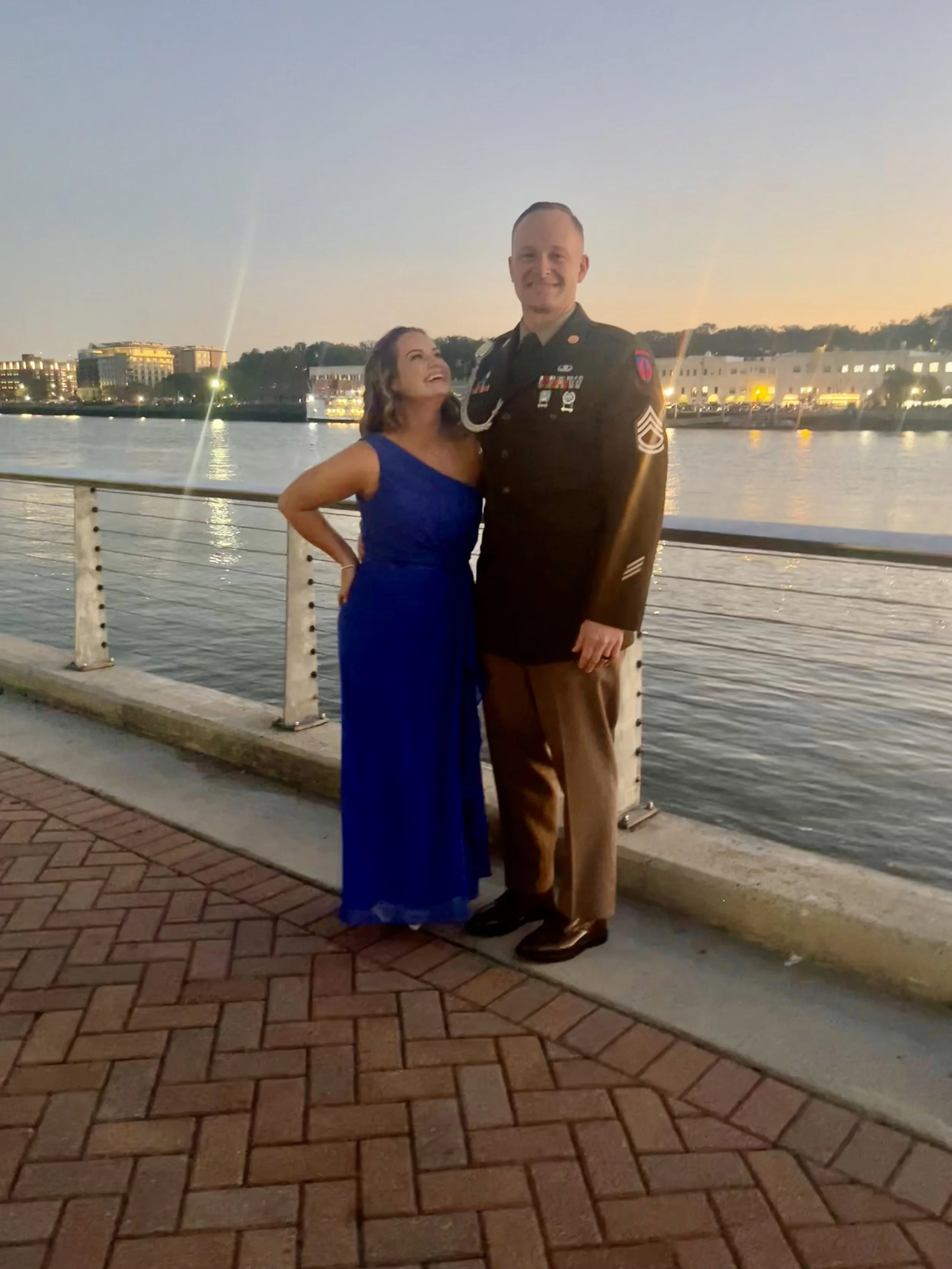 A woman in a blue evening gown and a man in a military uniform pose together by a waterfront at sunset.