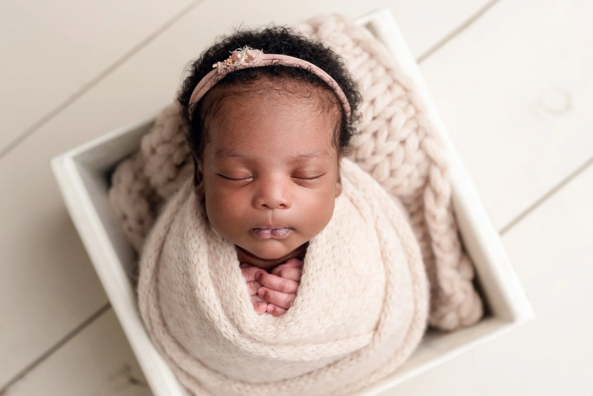 Newborn baby sleeping in a small white square box wrapped in light pink wrap wearing a dainty matching headband on a white wooden floor.