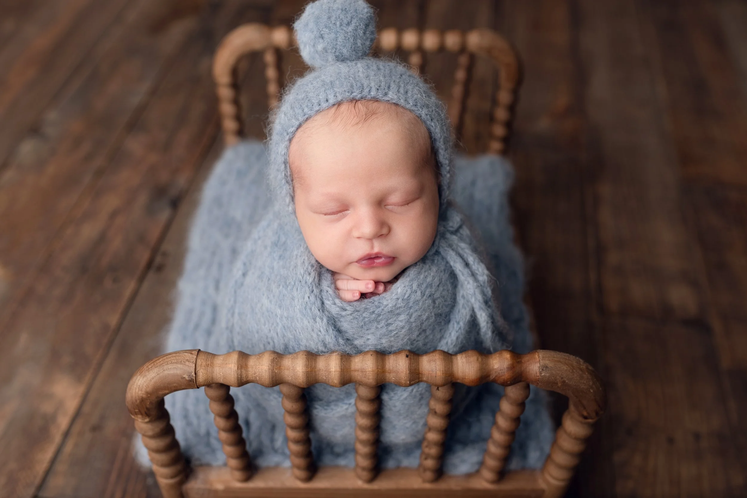Newborn baby wrapped in a light blue knitted wrap wearing a bonnet, sleeping on a small wooden spindle bed.