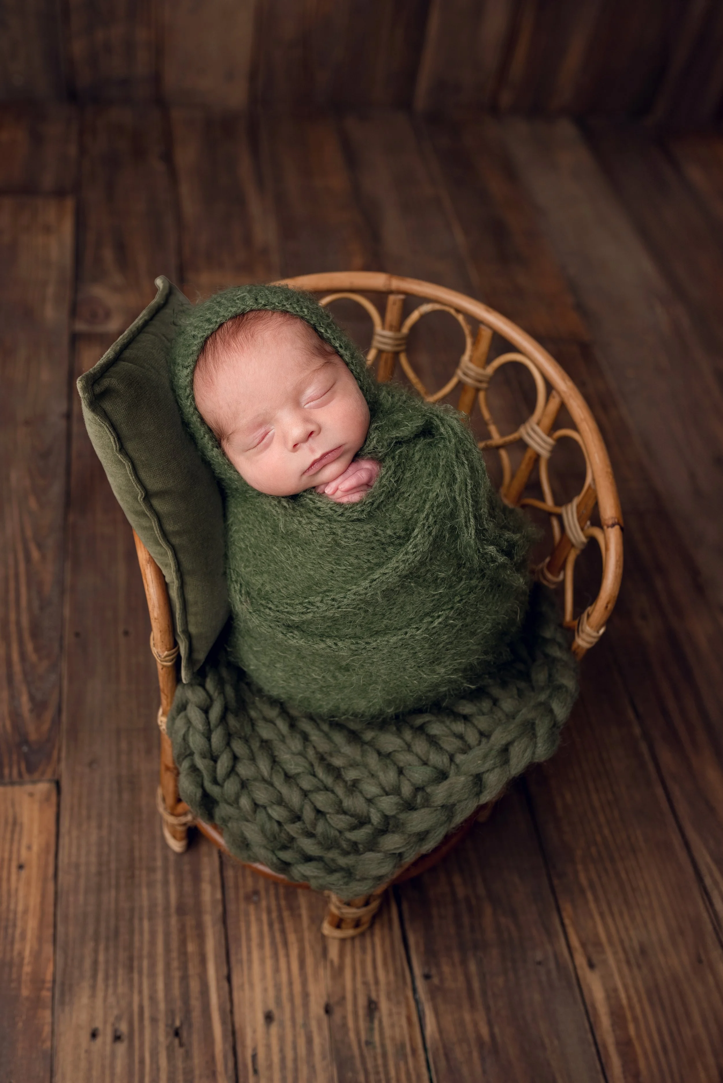 Sleeping newborn baby wrapped in a dark green wrap with matching bonnet on a small wooden chair on a dark brown wooden floor.