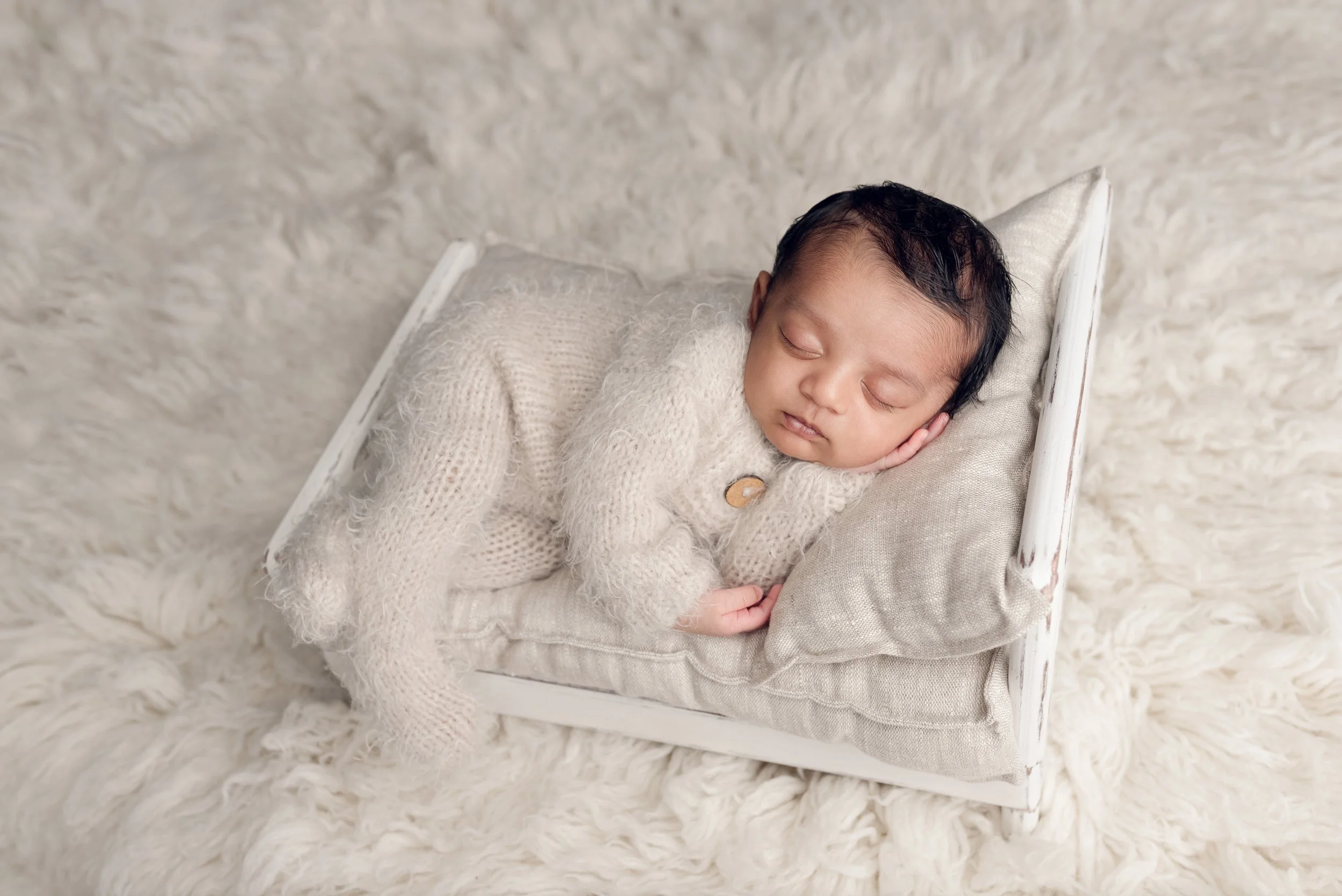 Newborn baby sleeping on a small white bed wearing a cream footed romper on a white fuzzy rug.