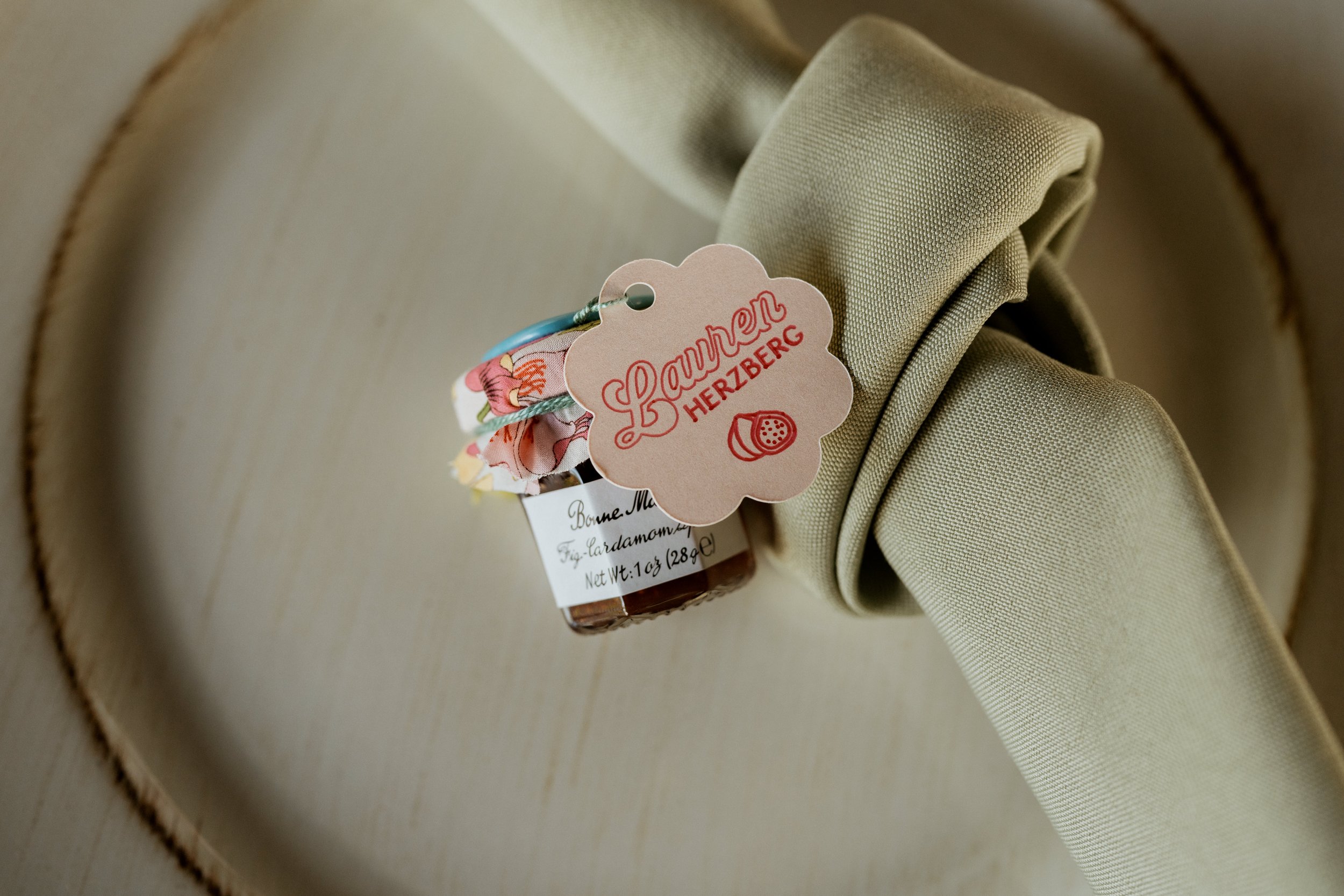 A small jar of Bonne Maman fig and cardamom jam with a floral cloth cover, attached to a beige napkin tied around a plate with a tag that reads "Launen HERZBERG."