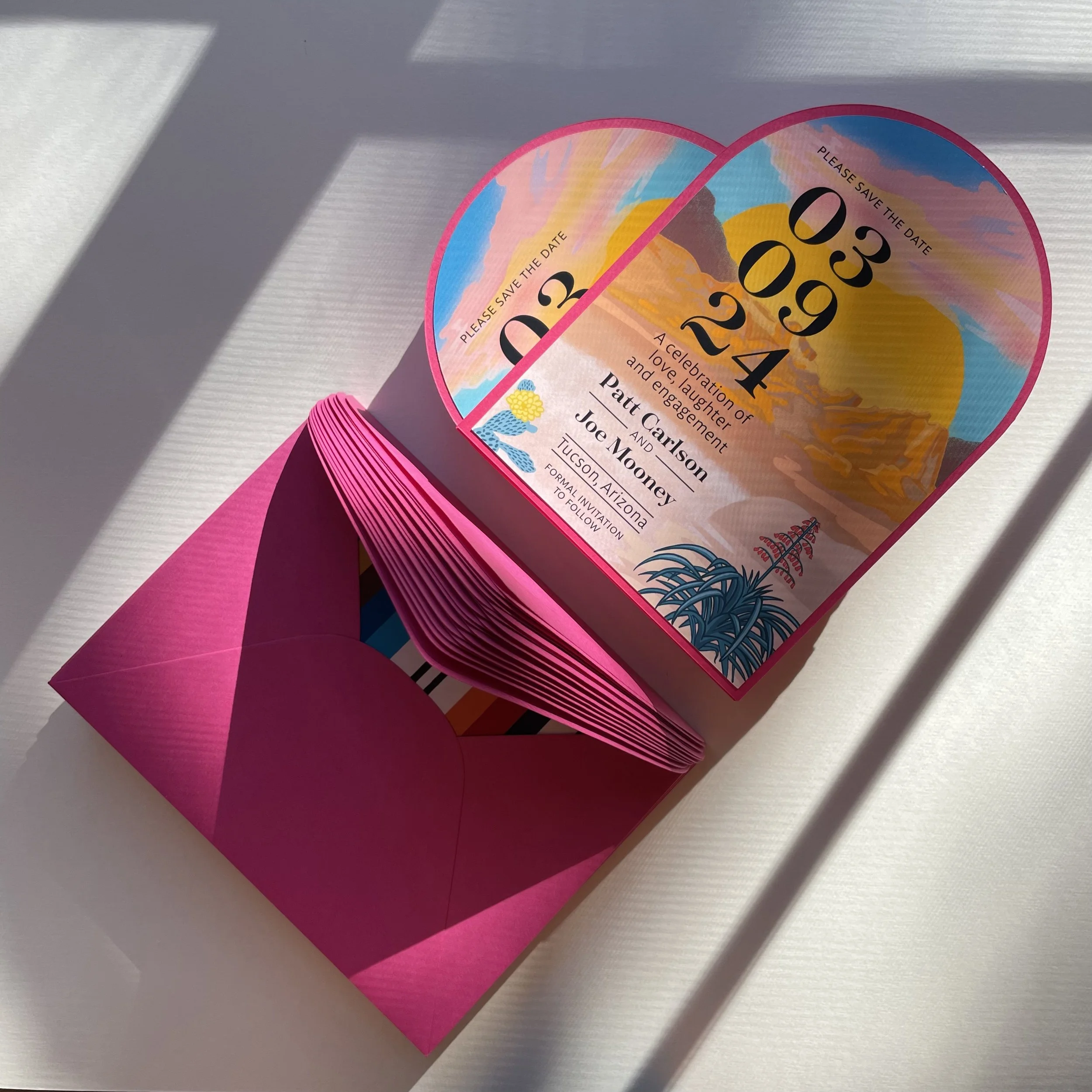 Colorful heart-shaped wedding invitation with sunset and mountain background, pink envelope, and floral accents.