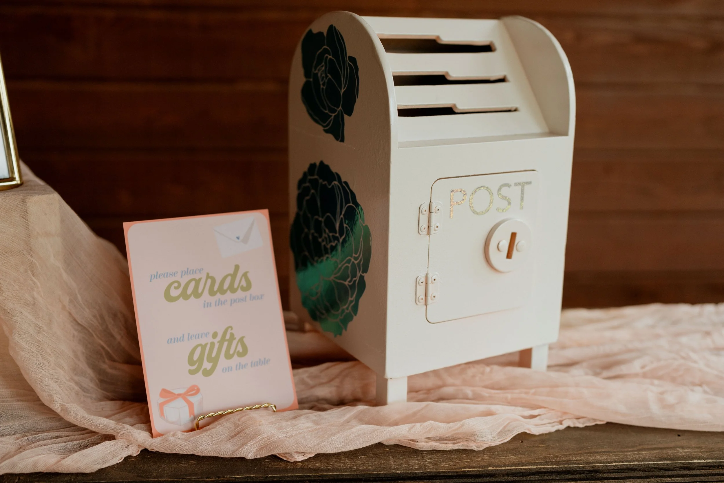 A white post box with black floral decals and a gold 'POST' label, placed on a wooden surface covered with pink fabric. Next to it, a pink sign instructs to place cards in the post box and leave gifts on the table.