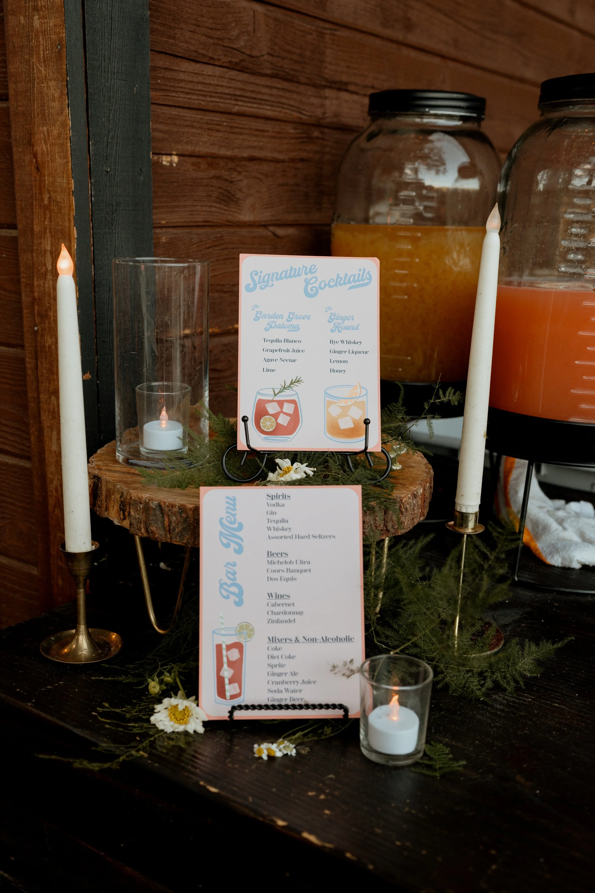 Wedding drink and menu setup with candles, rustic wood and floral decorations. Two large glass dispensers with beverages are behind the table, along with a small sign displaying signature cocktails and a menu listing spirits, beers, wines, and non-alcoholic mixers.