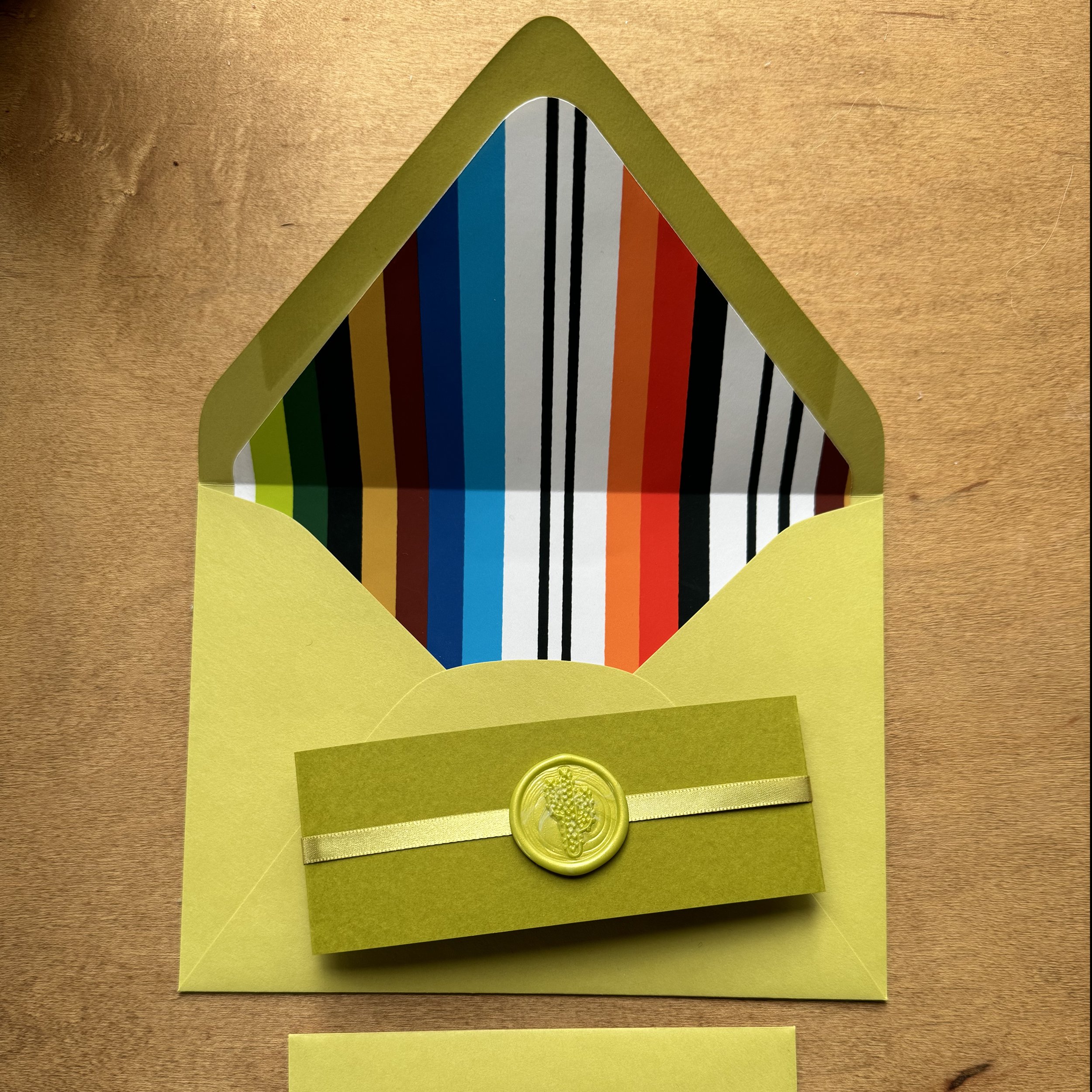 Yellow greeting card with a ribbon and wax seal, open to show colorful striped interior on a wooden surface.