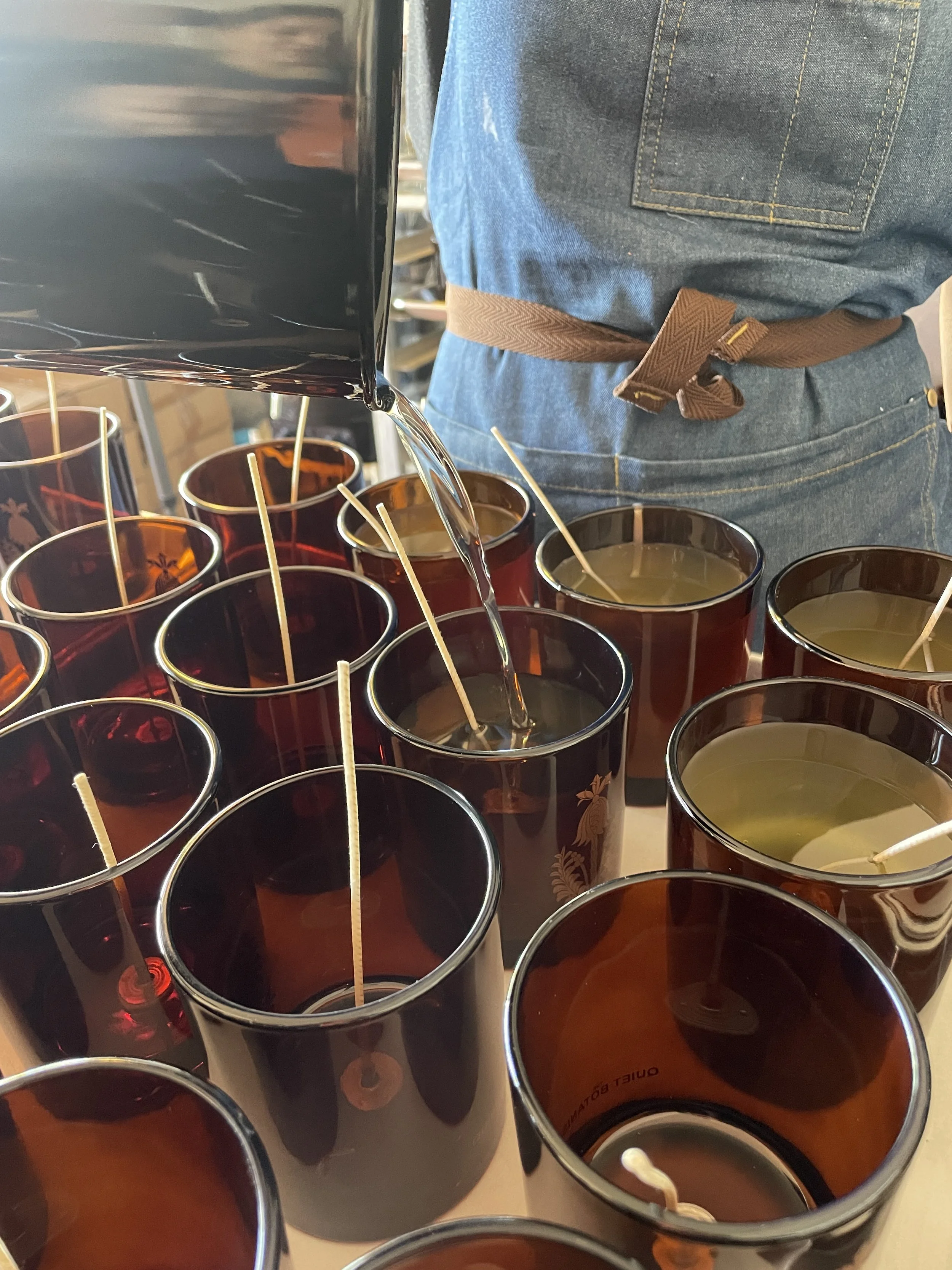 Close-up of a candlemaker with liquid above a small brown glass bottle, blurred bottles in the background.