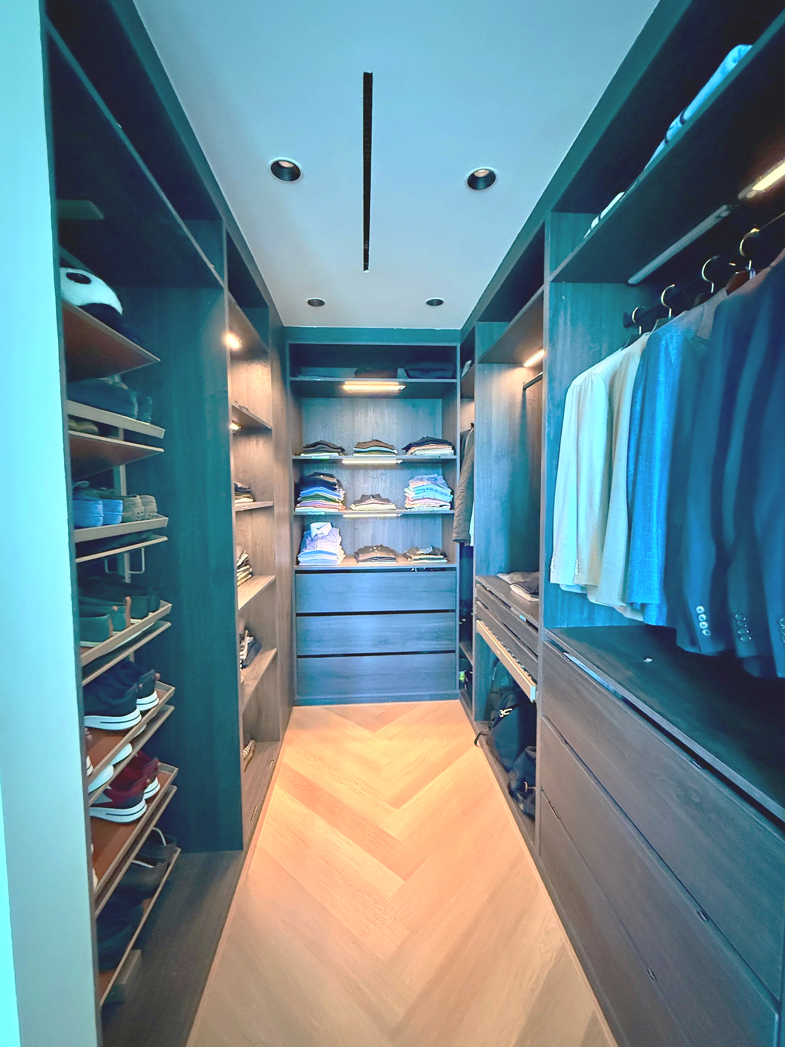 A walk-in closet with dark wood shelves and drawers, containing folded clothes, shoes, and hanging shirts.