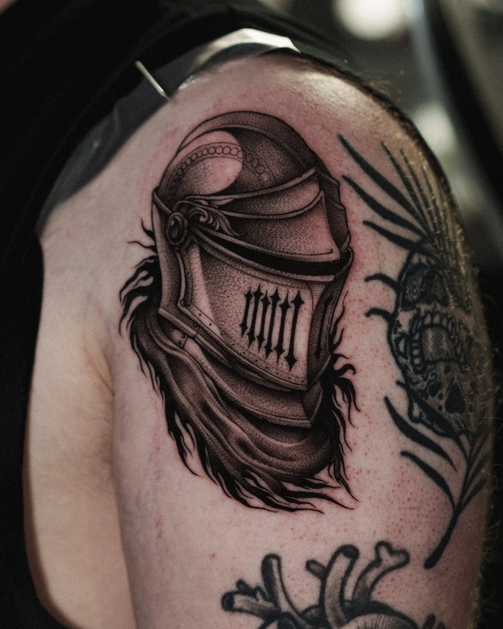 Close-up of a tattoo featuring a stylised, detailed medieval knights helmet with decorative elements surrounding it. Dark fantasy inspired tattoo by Jason James Smith