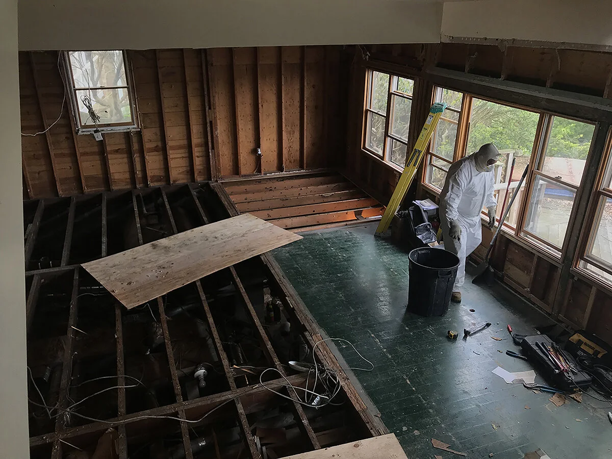 A person in protective suit cleaning a room under renovation. The room has large windows, with some wall panels removed, exposing wooden studs. The floor has a partially removed section, revealing the subfloor and pipes underneath. Construction tools
