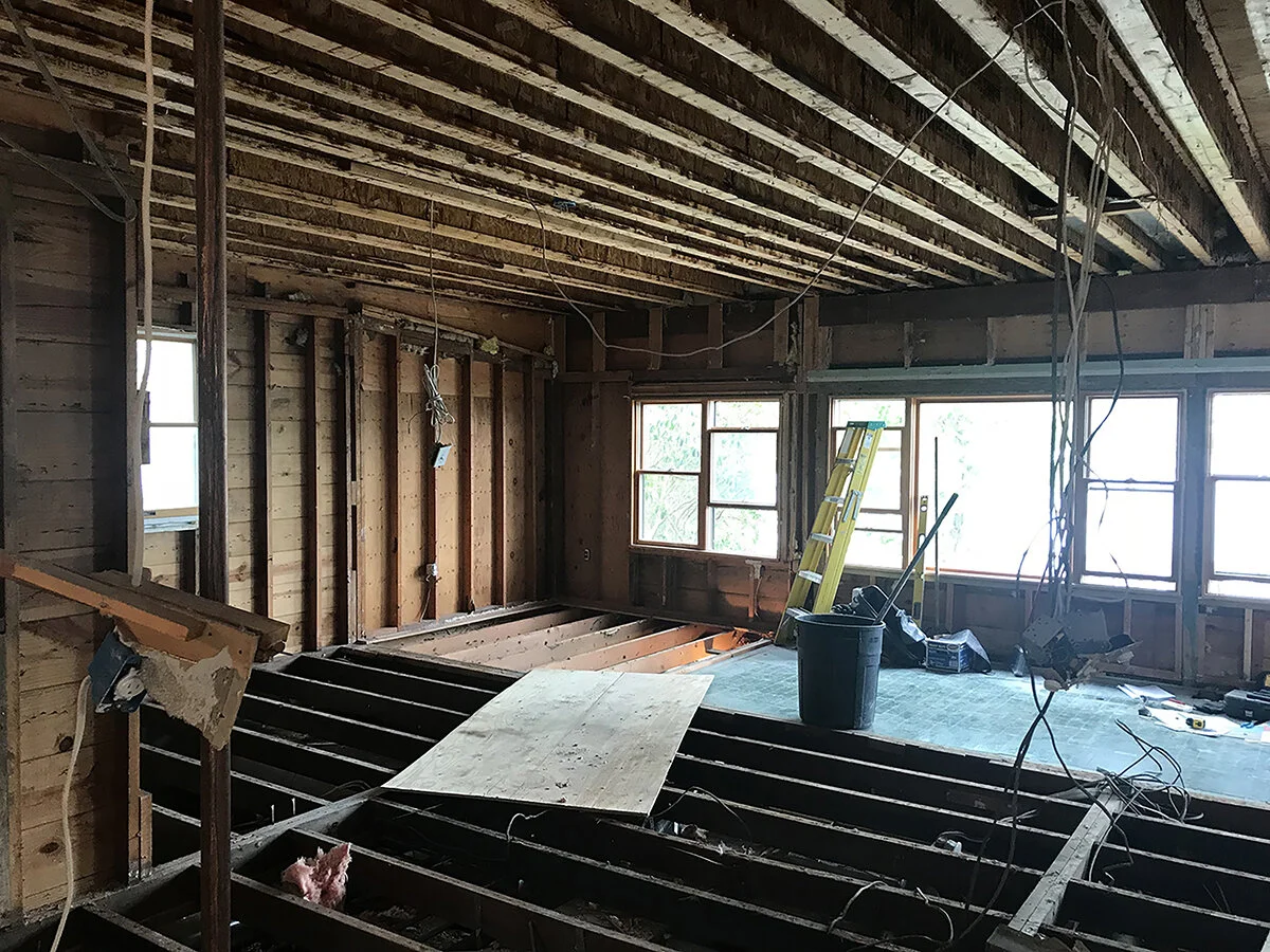 Interior of a house under renovation with exposed wooden beams, walls, and floors, construction tools, a ladder, and windows.