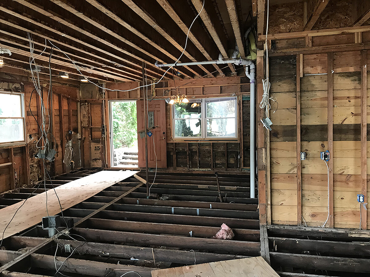 Interior of a house under renovation with exposed wooden studs, wiring hanging from the ceiling, and a partially removed floor, showing floor joists. There are a few windows and an open doorway leading outside.