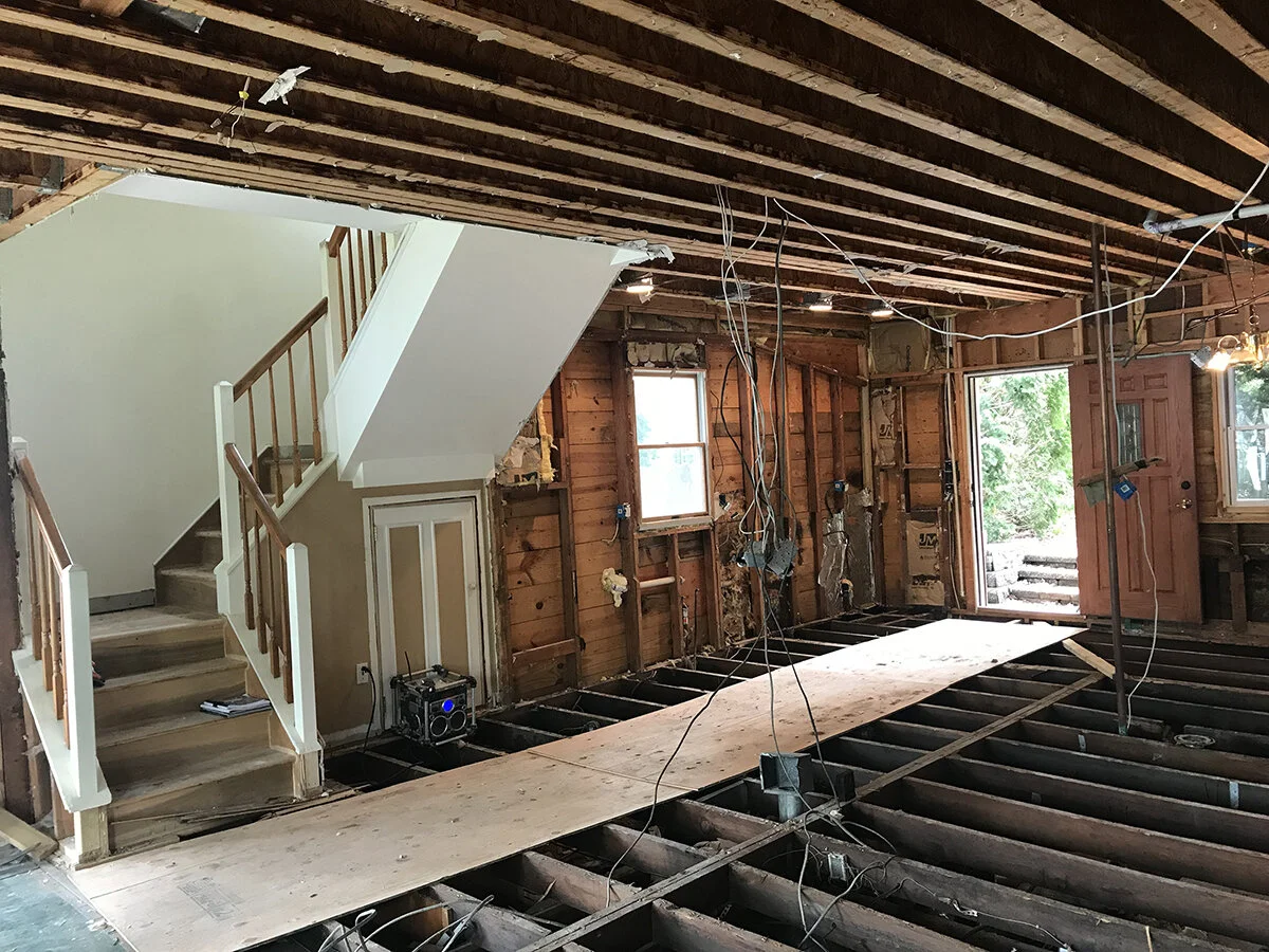 Interior of a house under renovation with exposed floor joists, wiring hanging from the ceiling, a staircase with a white railing, and an open front door leading outside.