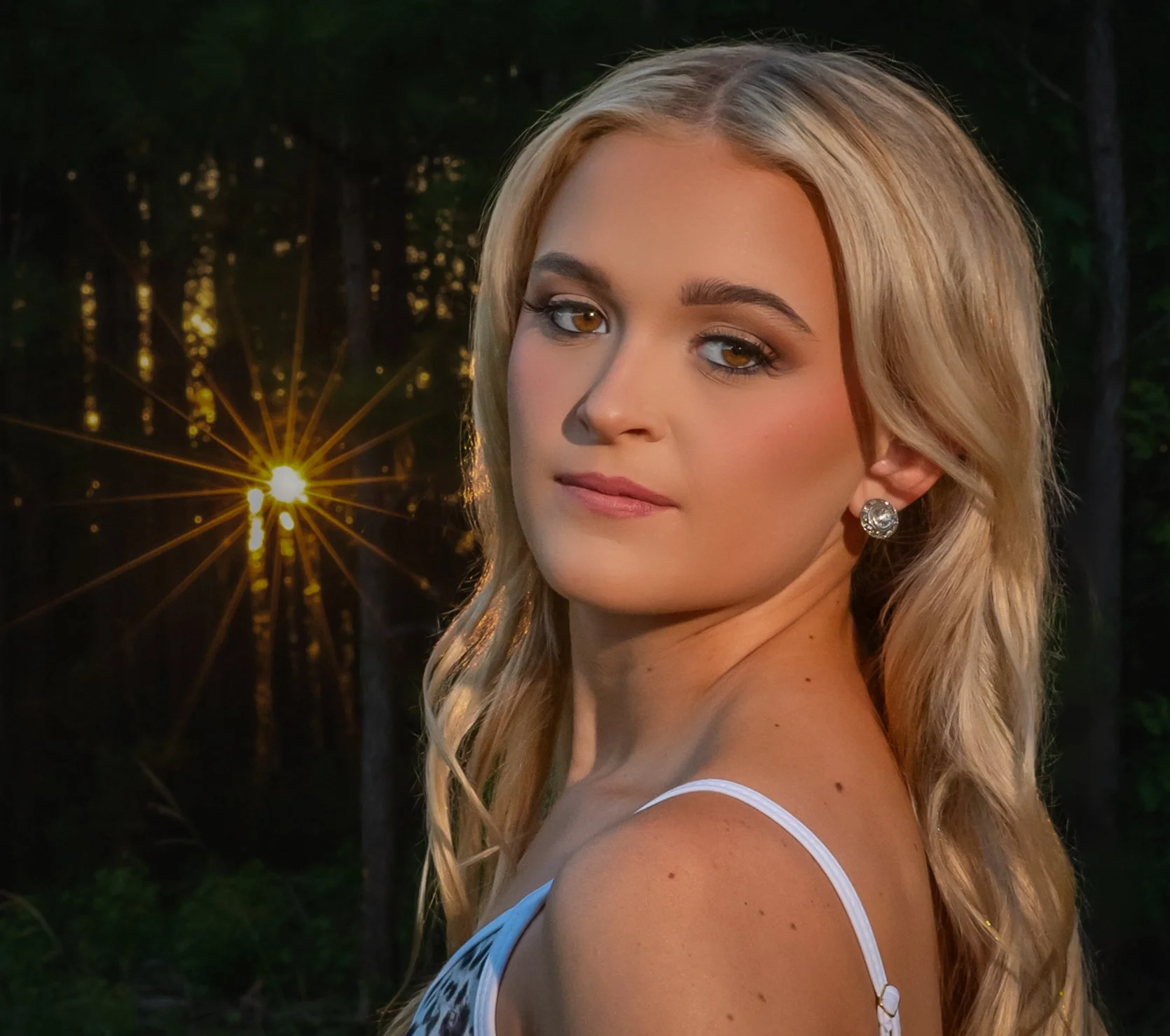 A woman with blonde hair and earrings outdoors during sunset or dusk, with trees and a bright light or star in the background.