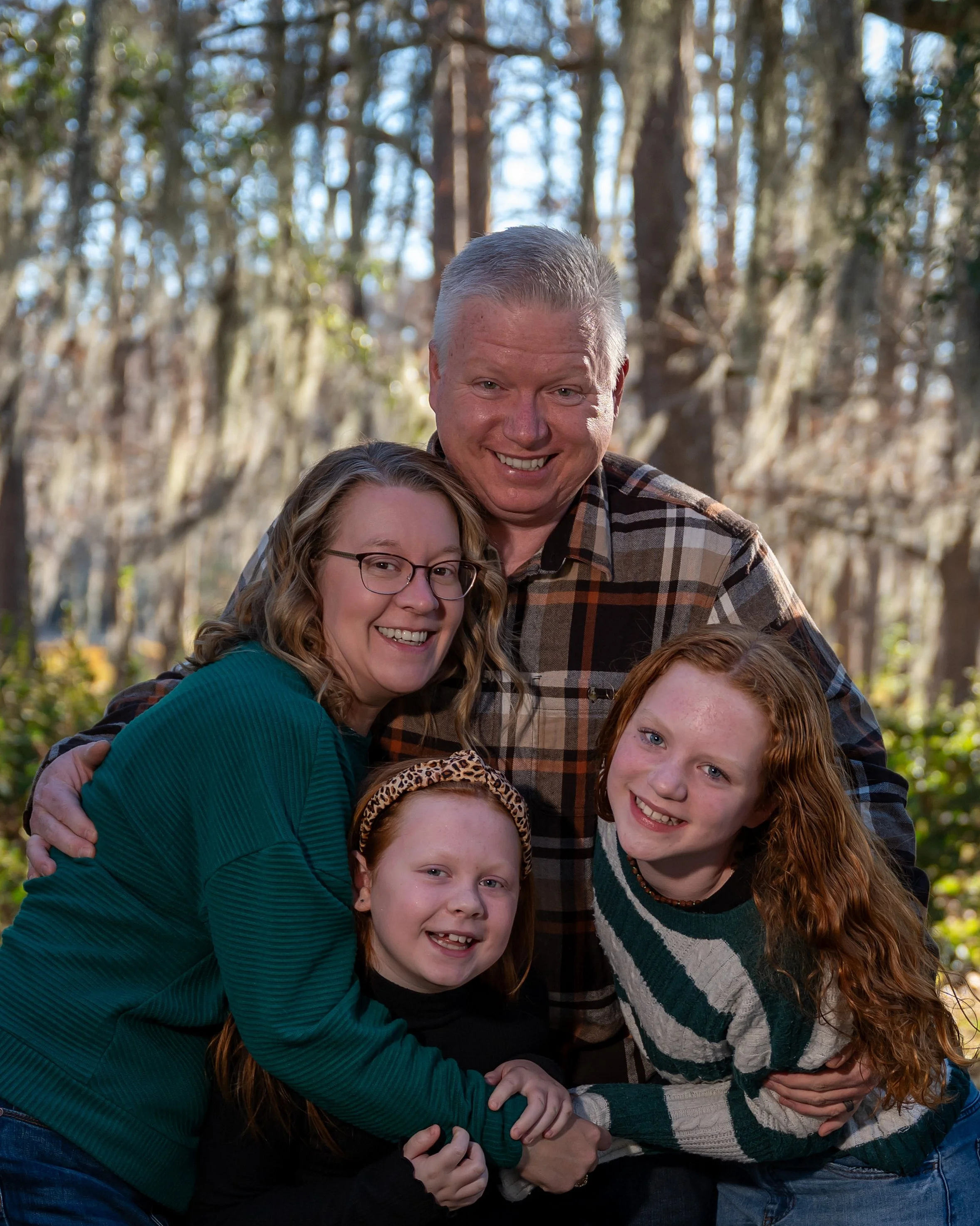 A group of five people standing outdoors in a wooded area, smiling and hugging each other. The group includes children and an adult man, all dressed in casual clothing.