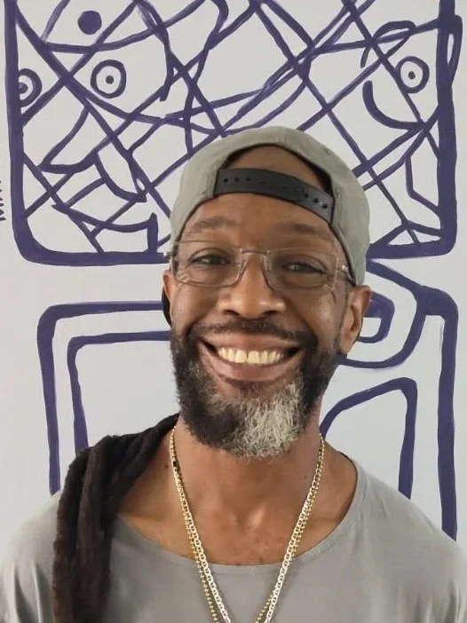 A smiling man with glasses, a beard with gray, wearing a backward baseball cap, gold chains, and a gray shirt, standing in front of a wall with abstract purple line art drawings.