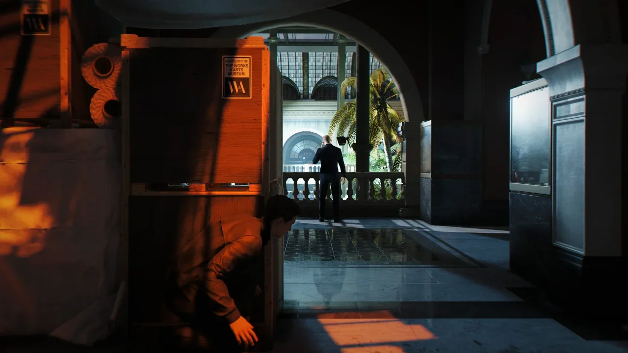 A stealth gameplay screenshot from 007 First Light showing a character crouching behind wooden crates in a museum interior to avoid being seen by a guard on a balcony.