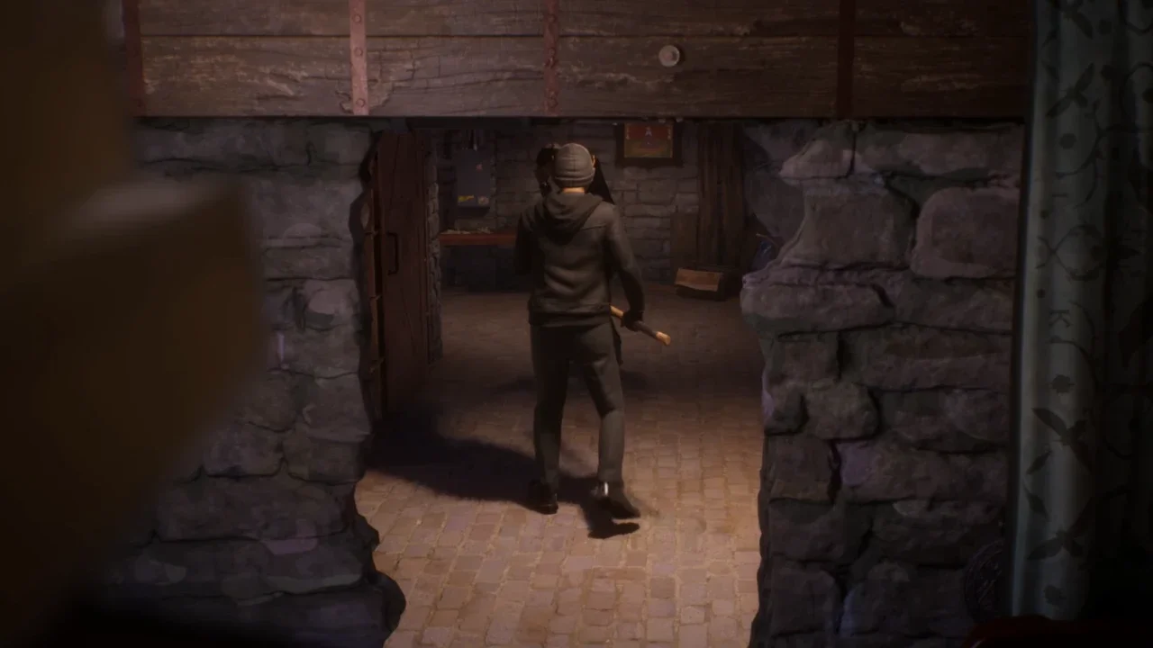 A character in a grey hoodie and beanie walks through a dimly lit stone cellar holding an axe in a gameplay screenshot from Life is Strange Reunion.
