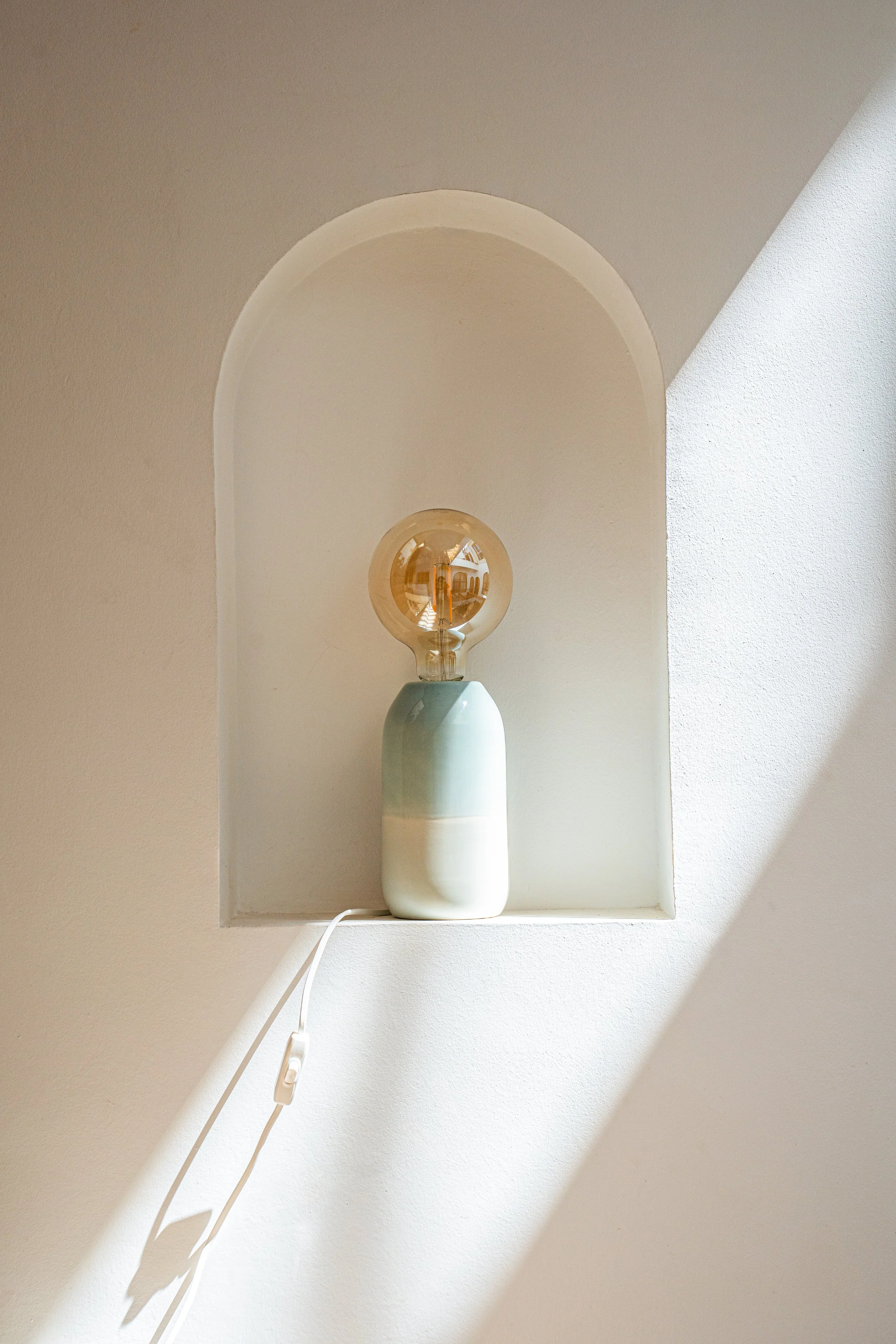 Ein Tablelampen-Design mit einem gezeichneten, transparenten Glühbirnenlampenschirm auf einem weißen Sockel, an einem Wandnischen mit viel natürlichem Licht.