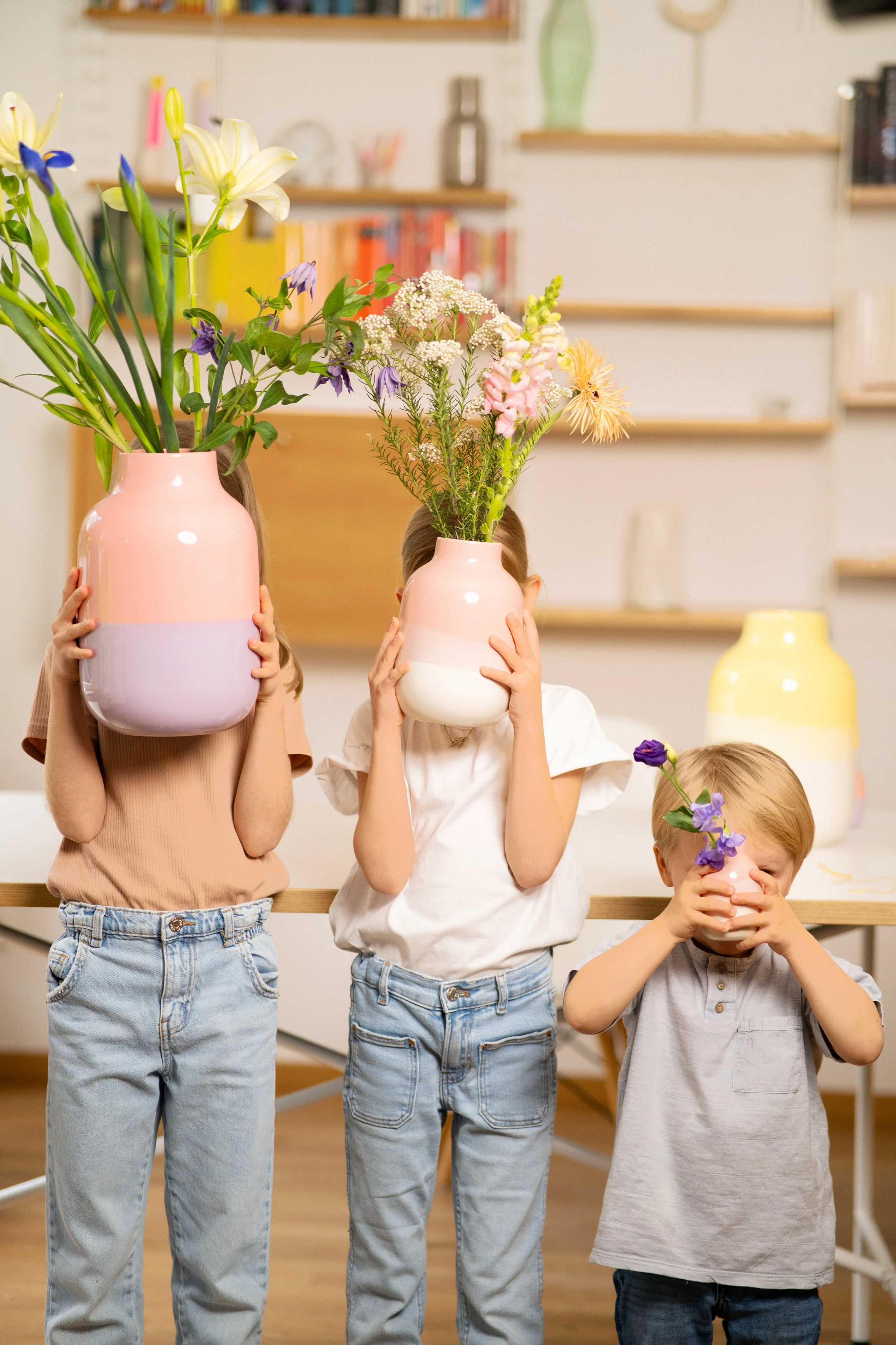 Drei Kinder halten große Vase mit Blumen vor Gesicht, im Hintergrund weitere Vasen mit Blumen in einem Raum mit Bücherregalen.