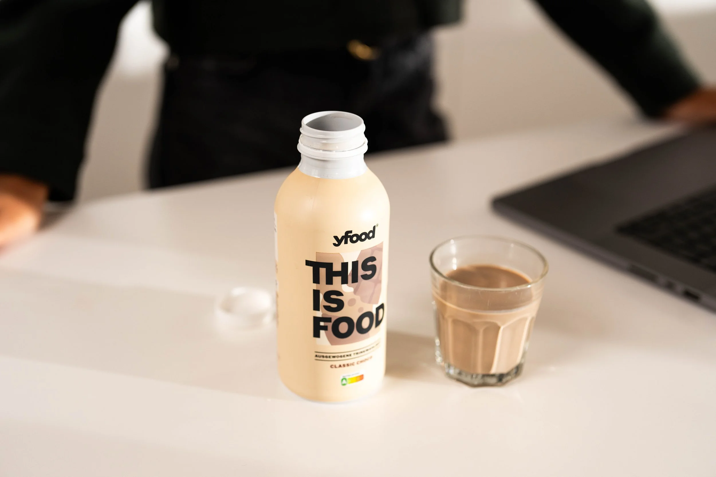 Eine Flasche mit der Aufschrift "THIS IS FOOD" neben einem Glas mit brauner Flüssigkeit auf einem weißen Tisch, im Hintergrund eine offene Laptop-Tastatur und eine Person in schwarzer Kleidung.