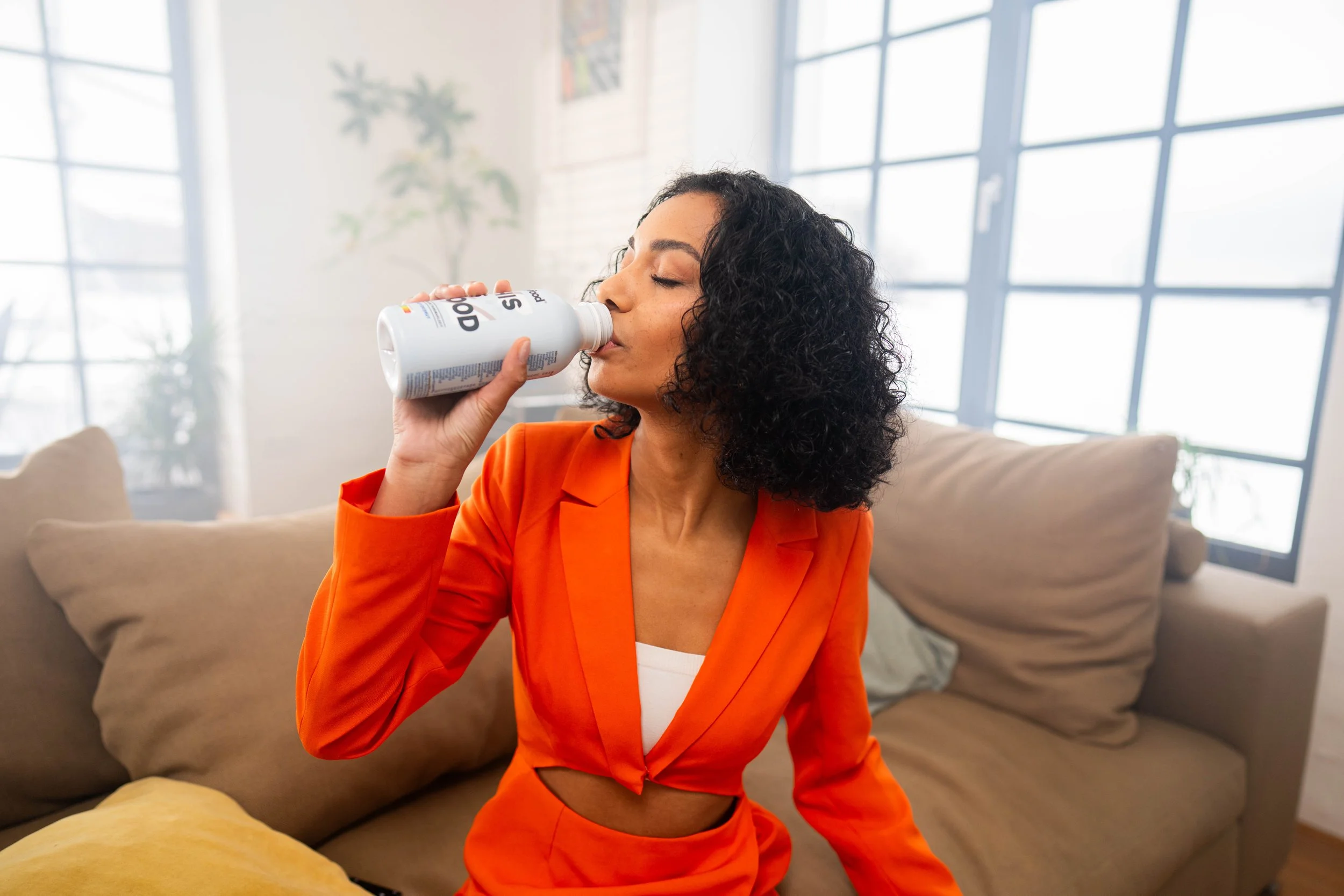 Frau in orange Kleidung trinkt aus einer Flasche in einem Wohnzimmer mit großen Fenstern.
