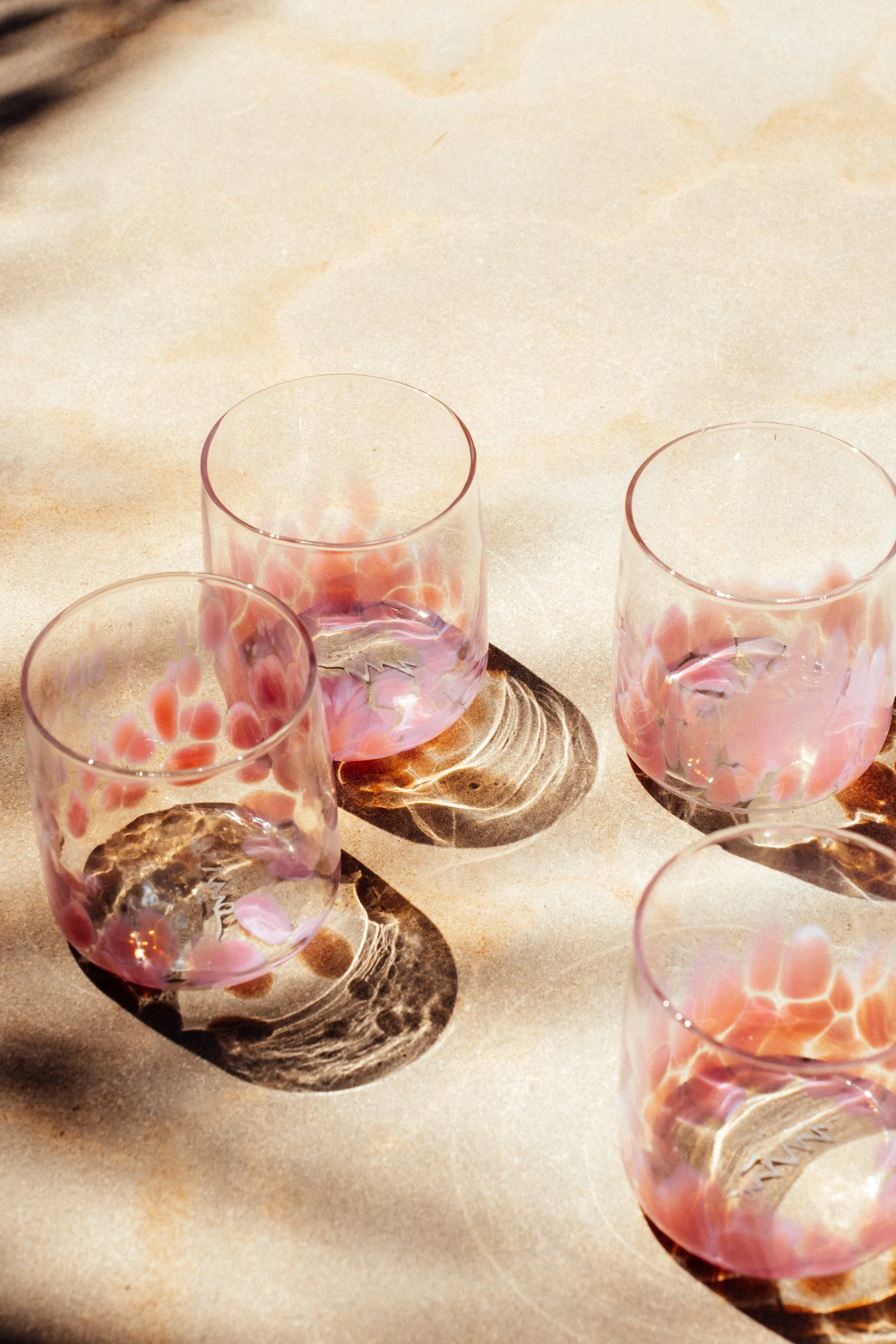 Vier klare Gläser mit pinkfarbenen Mustern, die Schatten auf einer hellen Oberfläche werfen.