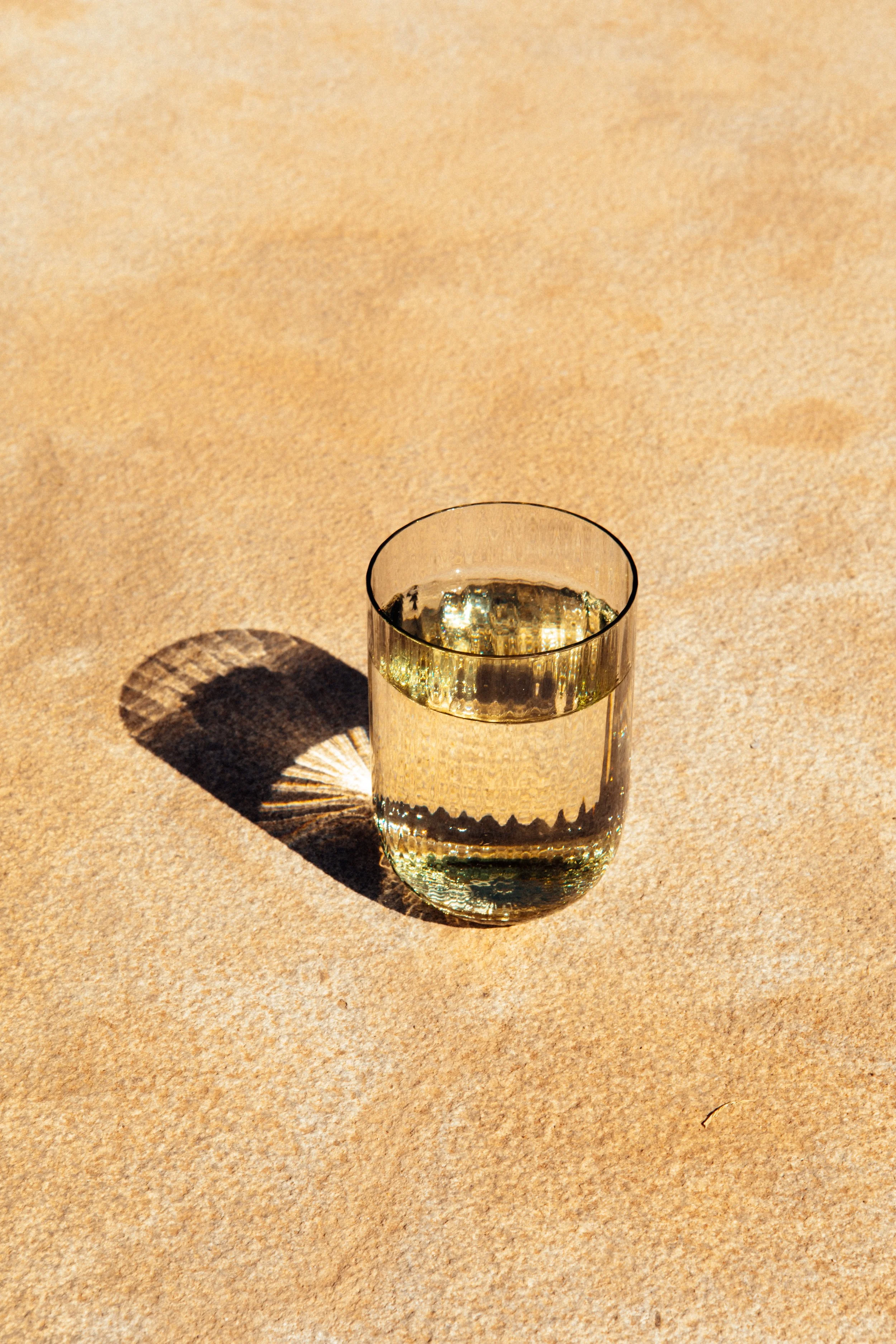 Ein Glas voller klaren, gelblichen Flüssigkeit, das auf sandigem Boden steht, mit einem langen Schatten, der durch das Sonnenlicht entsteht.