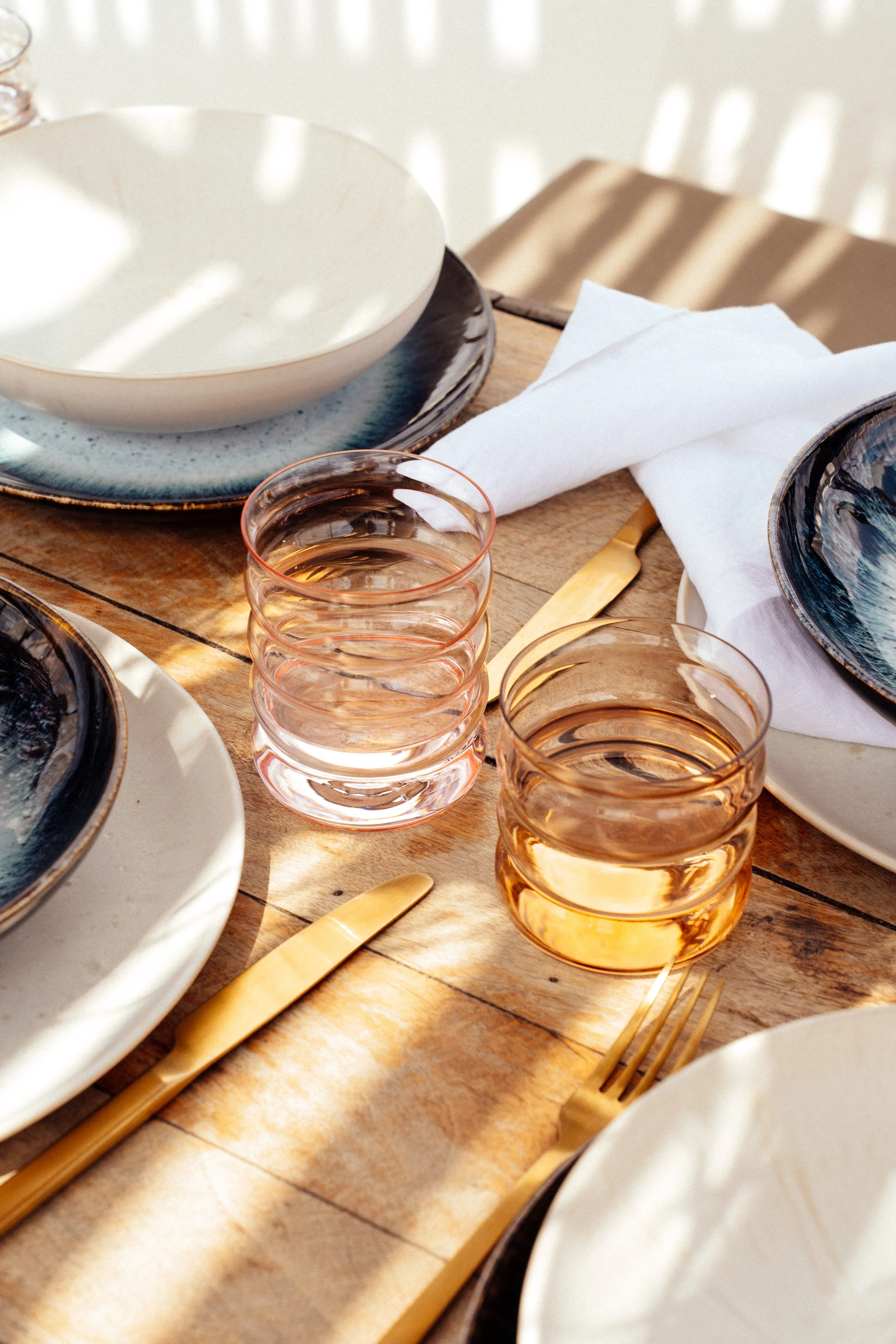 Tischgedeck mit bunten Tellern, goldenen Besteck und Gläsern auf holzmaserndem Tisch. Sonnenlicht wirft Schatten durch ein Gitter.