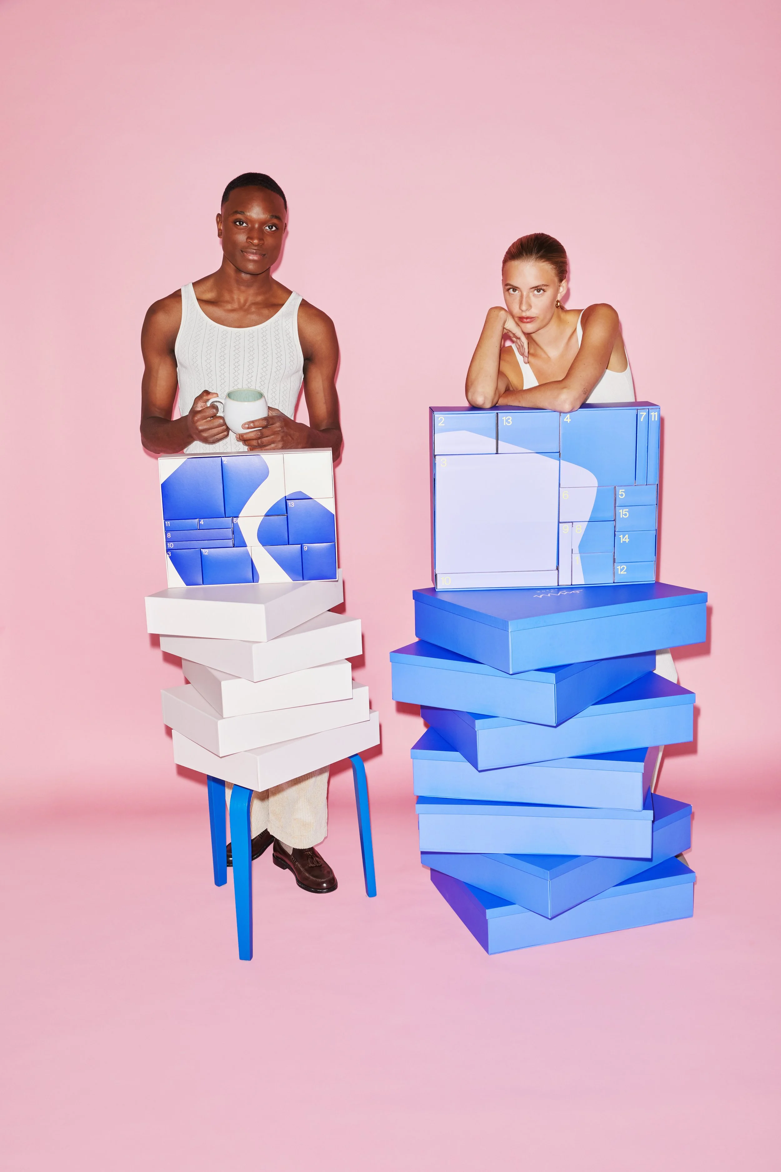 Zwei Frauen posieren vor einer pinken Hintergrund. Die Frauen stehen hinter großen blauen Kisten und Schachteln, die reichlich gestapelt sind. Die Frau auf der linken Seite hält eine Tasse, während die Frau auf der rechten Seite ihre Arme auf die Kis