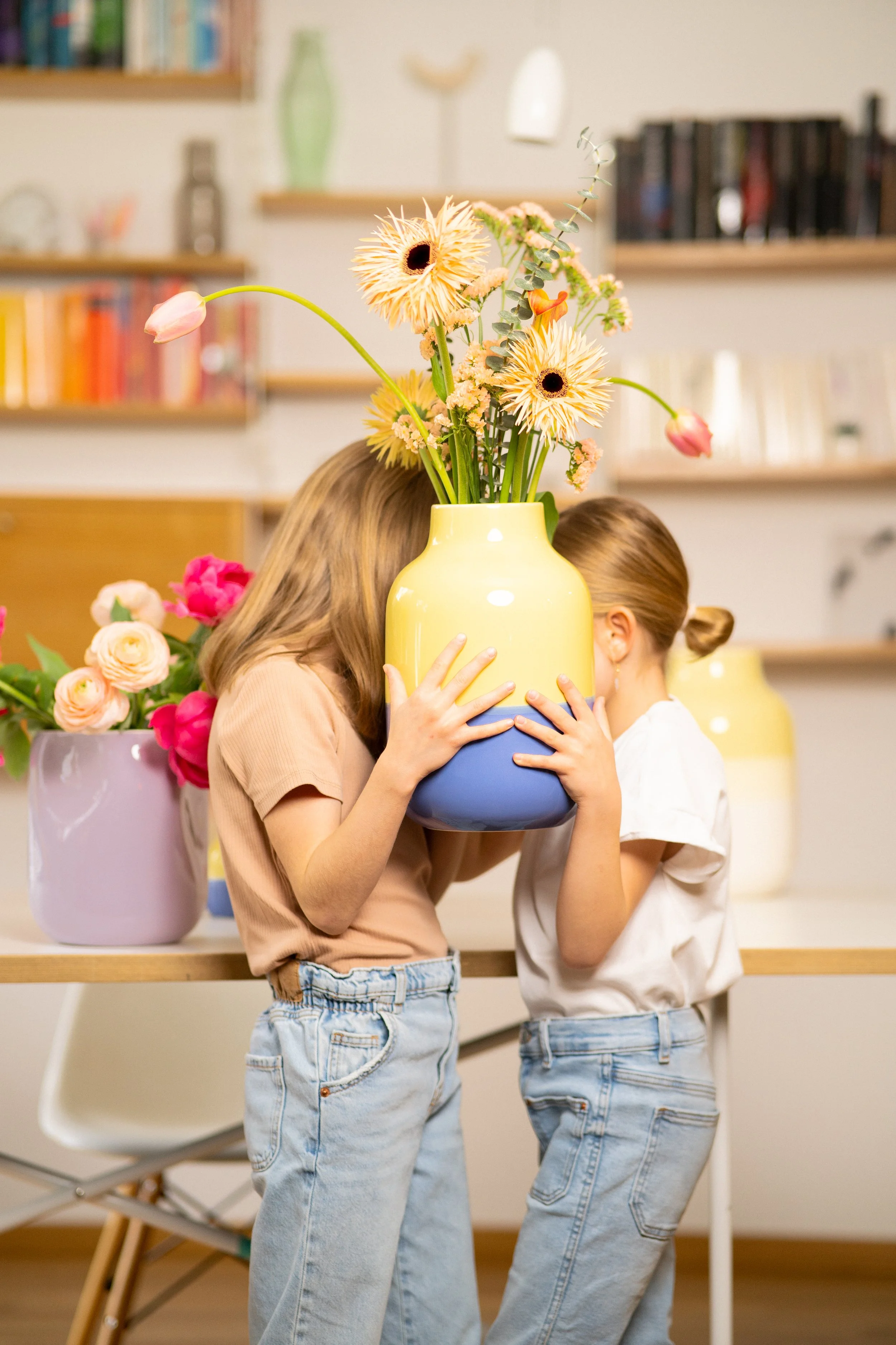 Zwei Kinder verstecken sich hinter einer großen bunten Vase mit Blumen in einem Wohnzimmer.