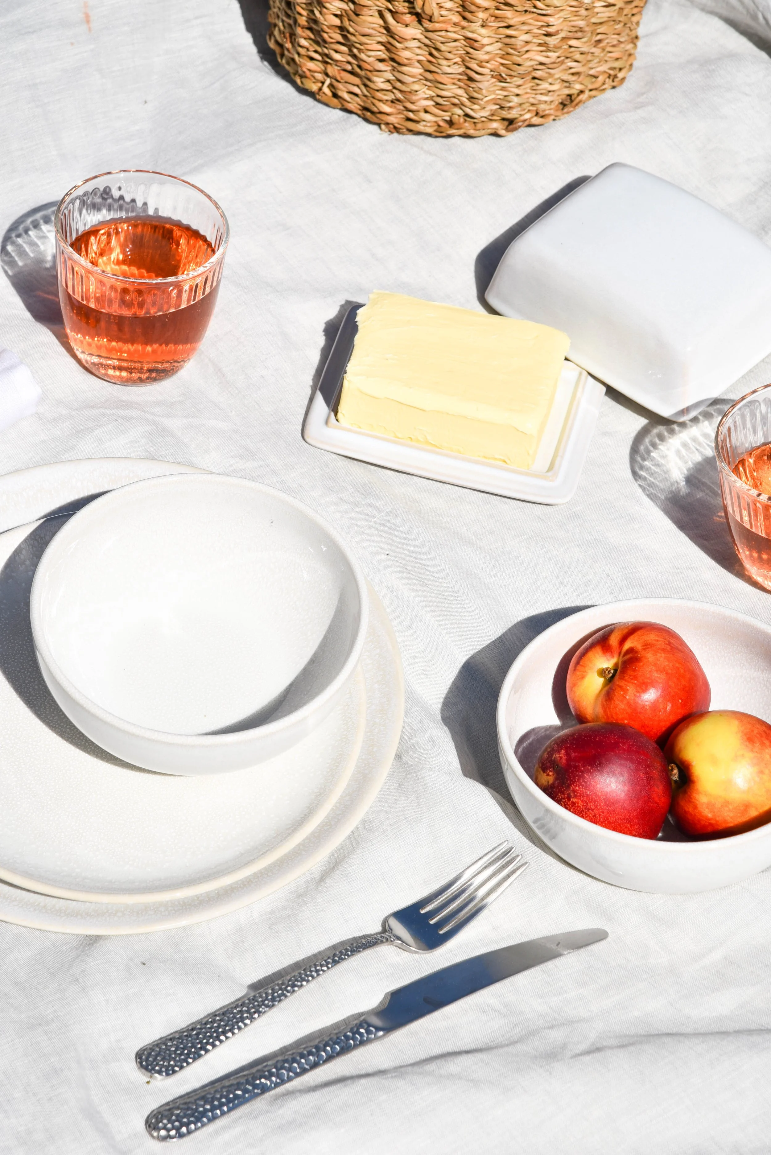 Ein gedeckter Tisch mit weißen Tellern, einem Messer und einer Gabel, einer Schüssel mit Äpfeln, einer Schale mit Butter, einem Stück Käse, einem Glas Roséwein und einem weißen Butterbrotpapier.